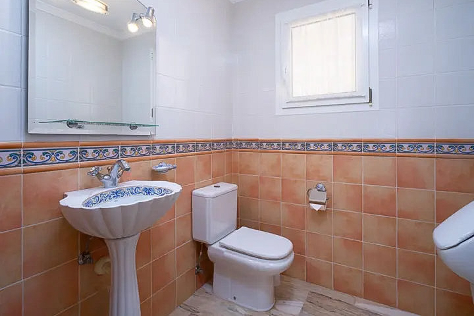 Gastentoilet met gedecoreerde wastafel op zuil, terracotta en witte wandtegels met sierstrip, grote spiegel en een klein raam voor natuurlijk licht.