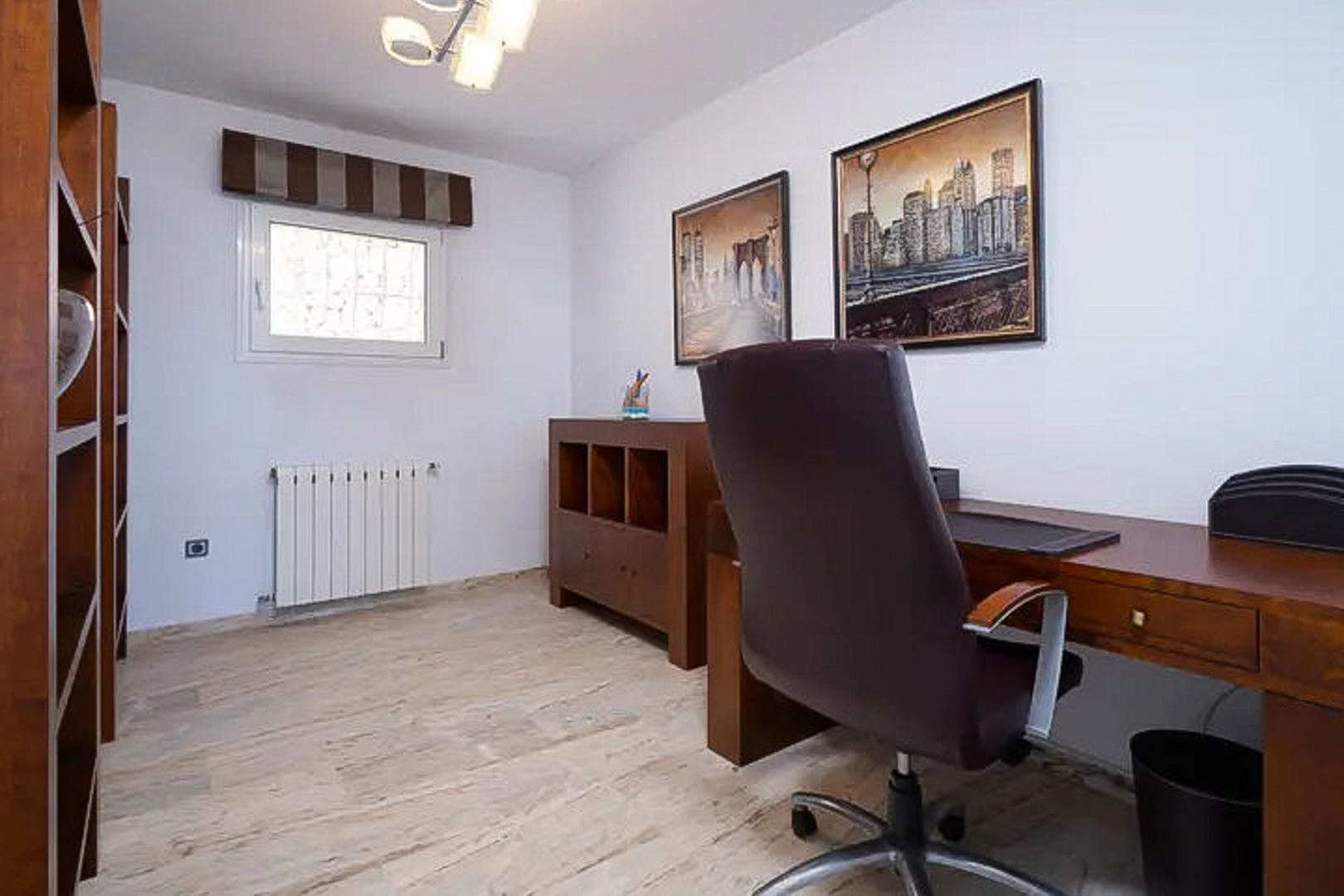 Licht thuiskantoor met houten meubilair, een functioneel bureau en een lederen stoel. Voorzien van kasten, een lichte stenen vloer en decoratieve stadsgezichten.