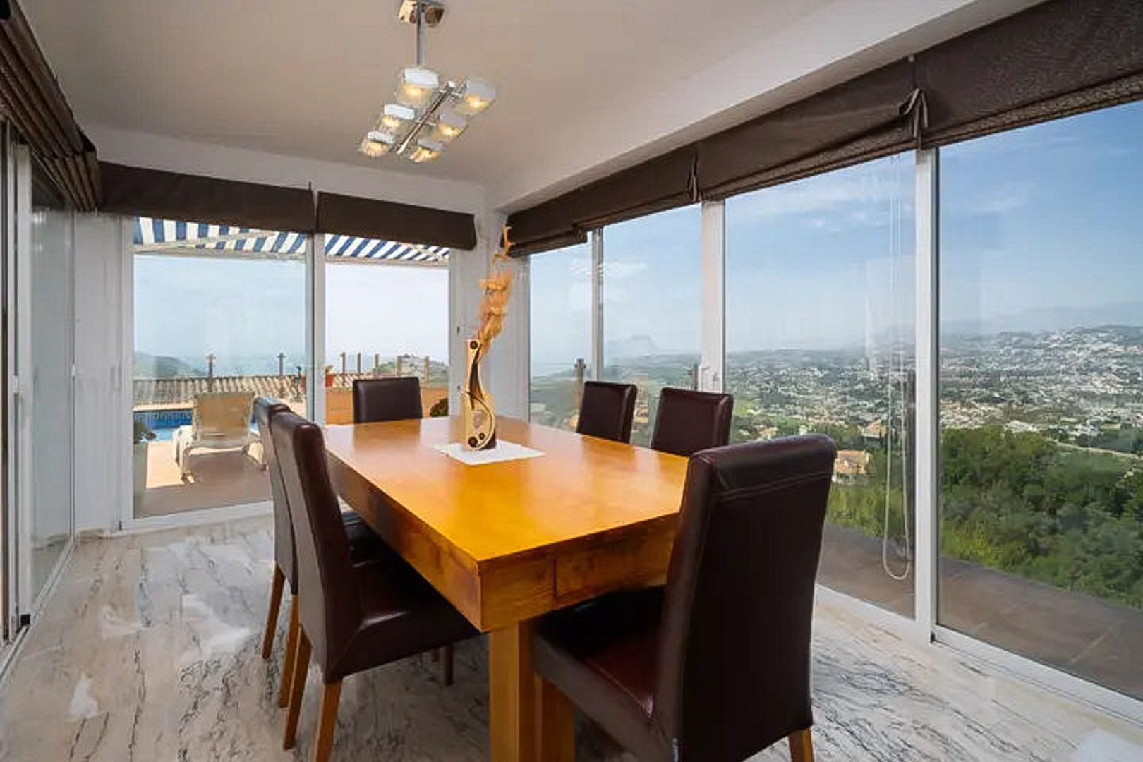 Lichte eetkamer met massief houten tafel en lederen stoelen, omgeven door grote glazen schuifdeuren met panoramisch uitzicht op zee en toegang tot het terras.