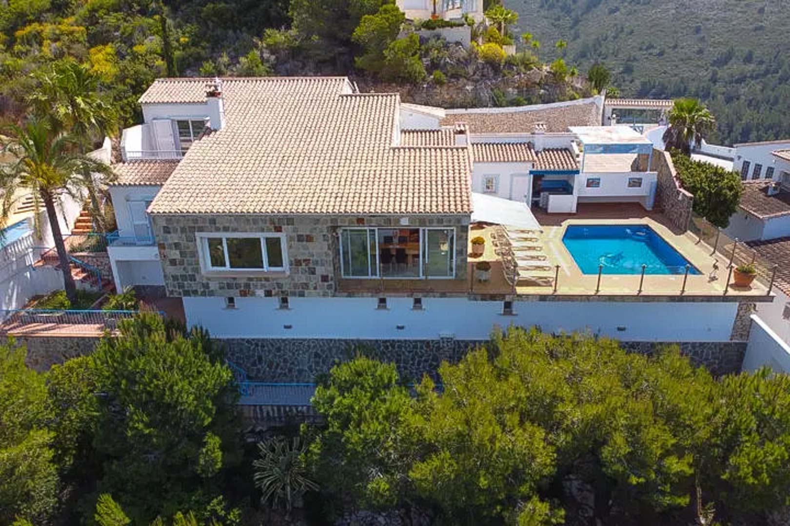 Luchtfoto van de villa met natuurstenen muren, pannendaken en een verhoogd terras met privézwembad en ligweide tussen de pijnbomen.