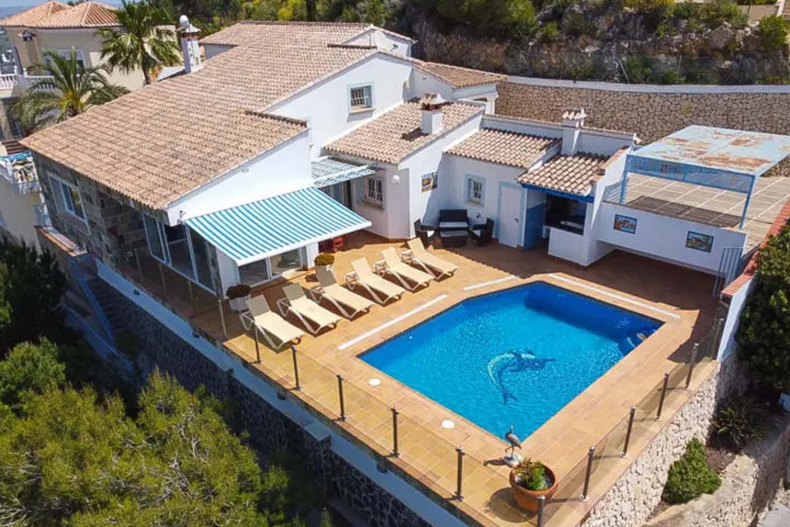 Luchtfoto van de villa met privézwembad, terras met ligbedden, gestreept zonnescherm en buitenbar, afgewerkt met glazen balustrades.