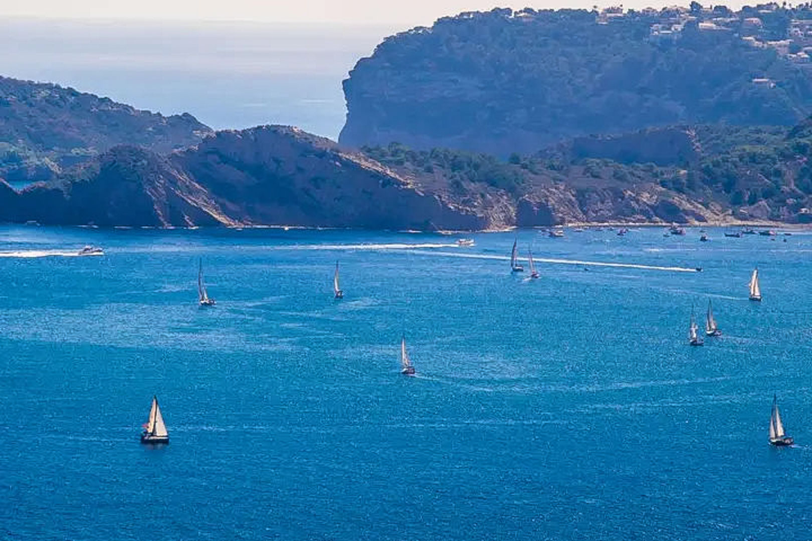 Panoramisch uitzicht op de Middellandse Zee in Moraira, met zeilboten die langs de rotsachtige kustlijn van de Costa Blanca varen.