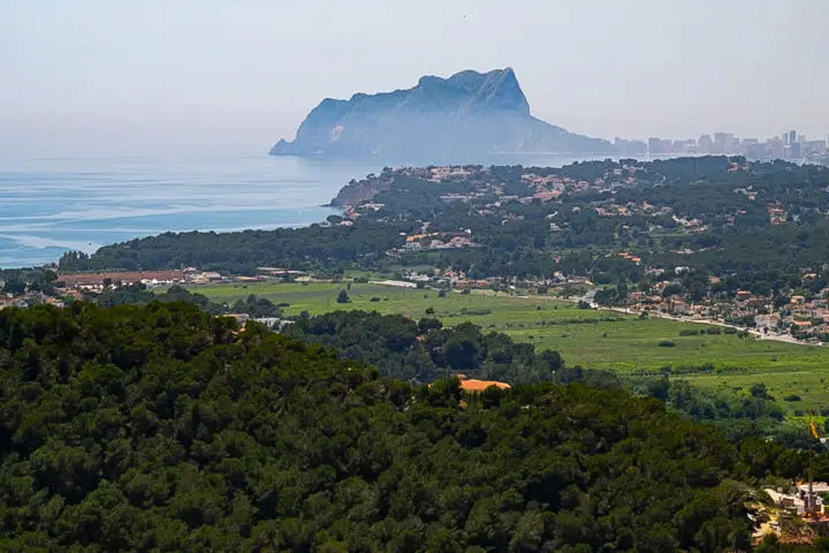 Panoramisch uitzicht op de Peñón de Ifach en de kustlijn, met het contrast tussen de dichte dennenbossen en het blauw van de Middellandse Zee.