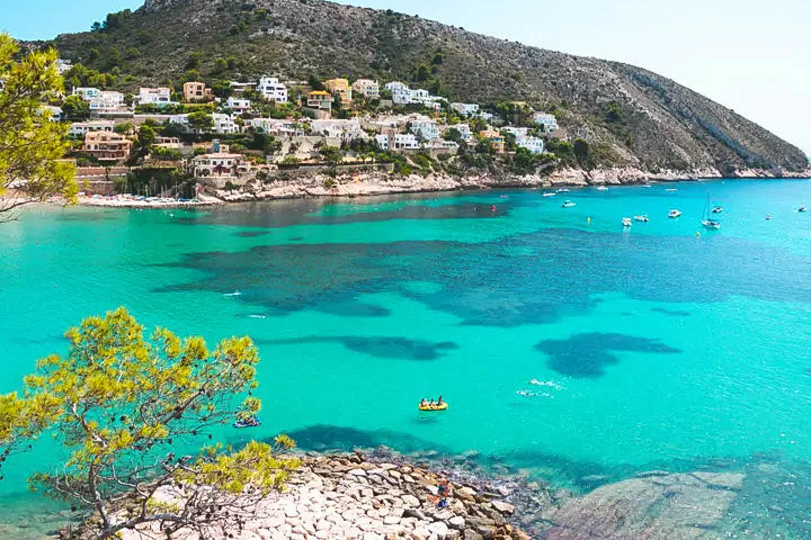 Panoramisch uitzicht op een baai in Moraira met kristalhelder turquoise water, mediterrane pijnbomen en witte villa's tegen de berghelling.