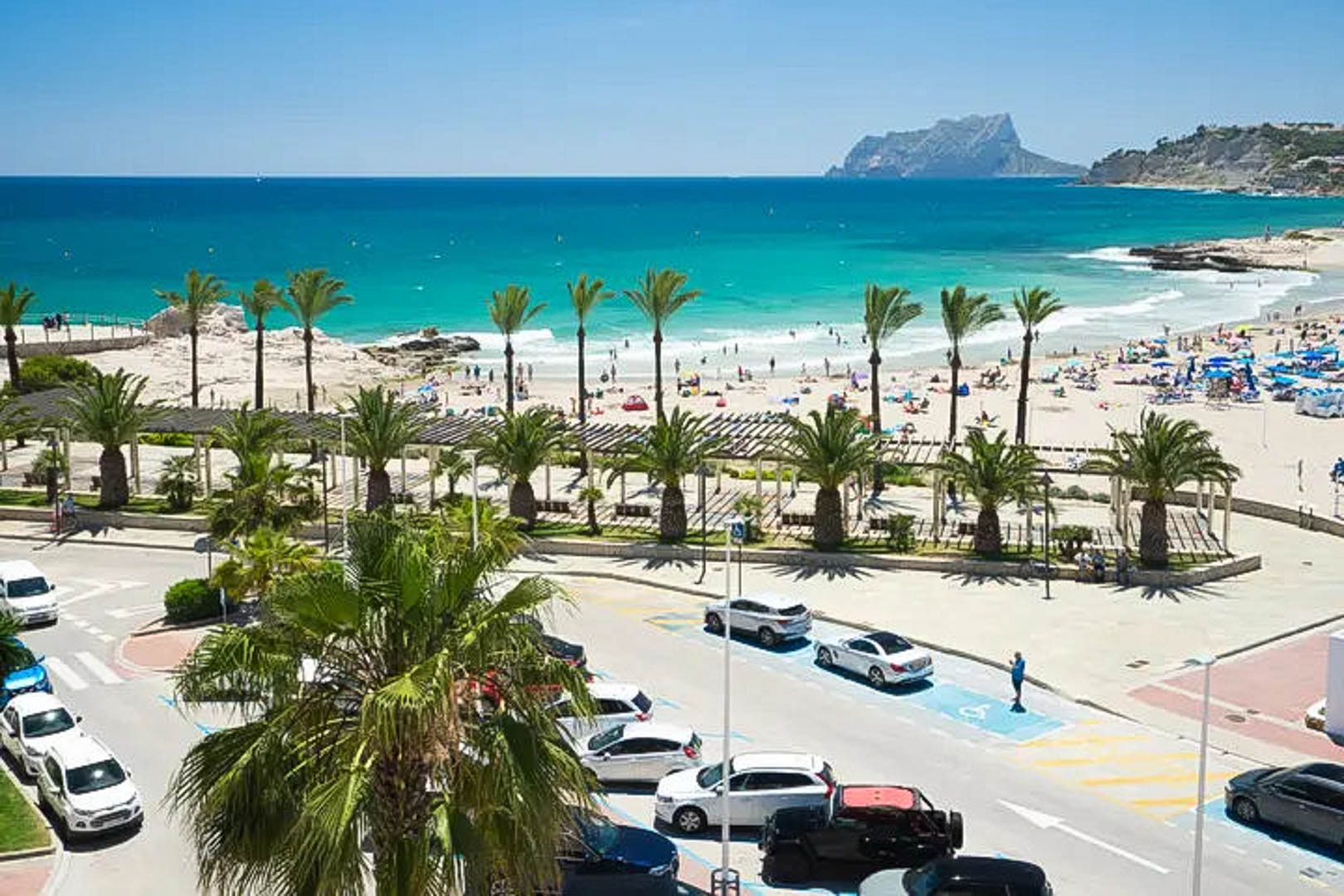 Panoramisch uitzicht op het zandstrand van Moraira met turquoise water, de Peñón de Ifach op de achtergrond en een promenade met palmbomen.