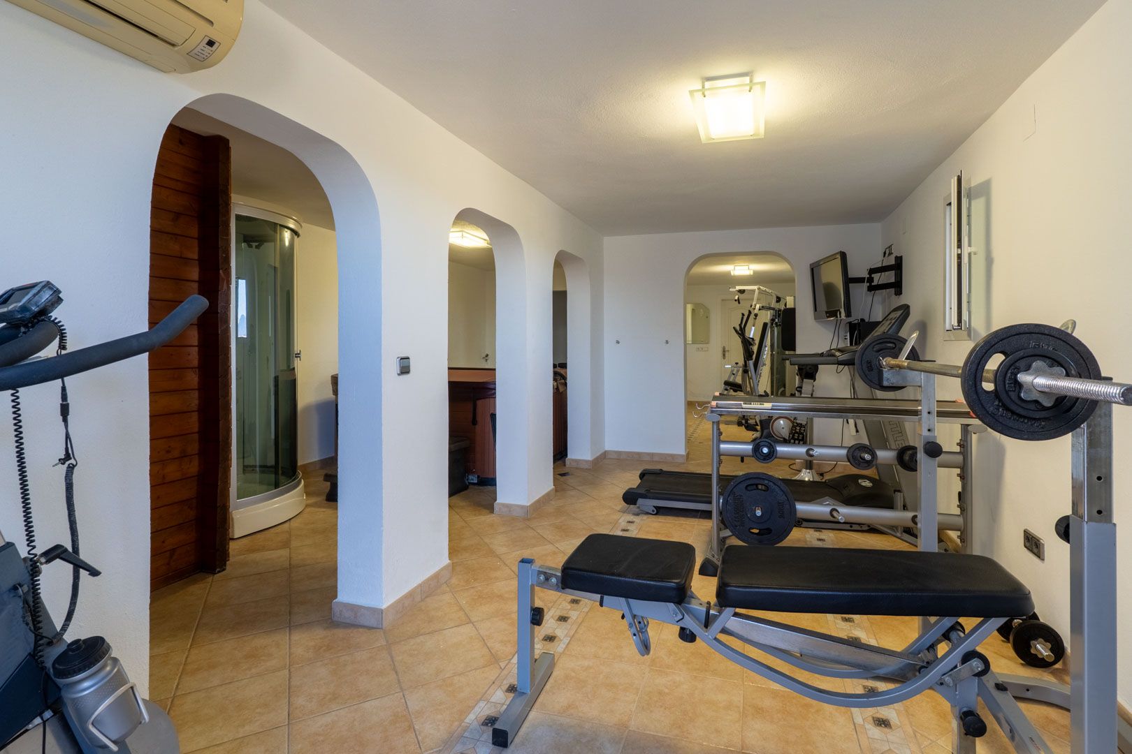 Privé-fitnessruimte en wellnessruimte met krachttrainingstoestellen, loopband en gewelfde doorgangen naar de sauna, stoomdouche en jacuzzi.