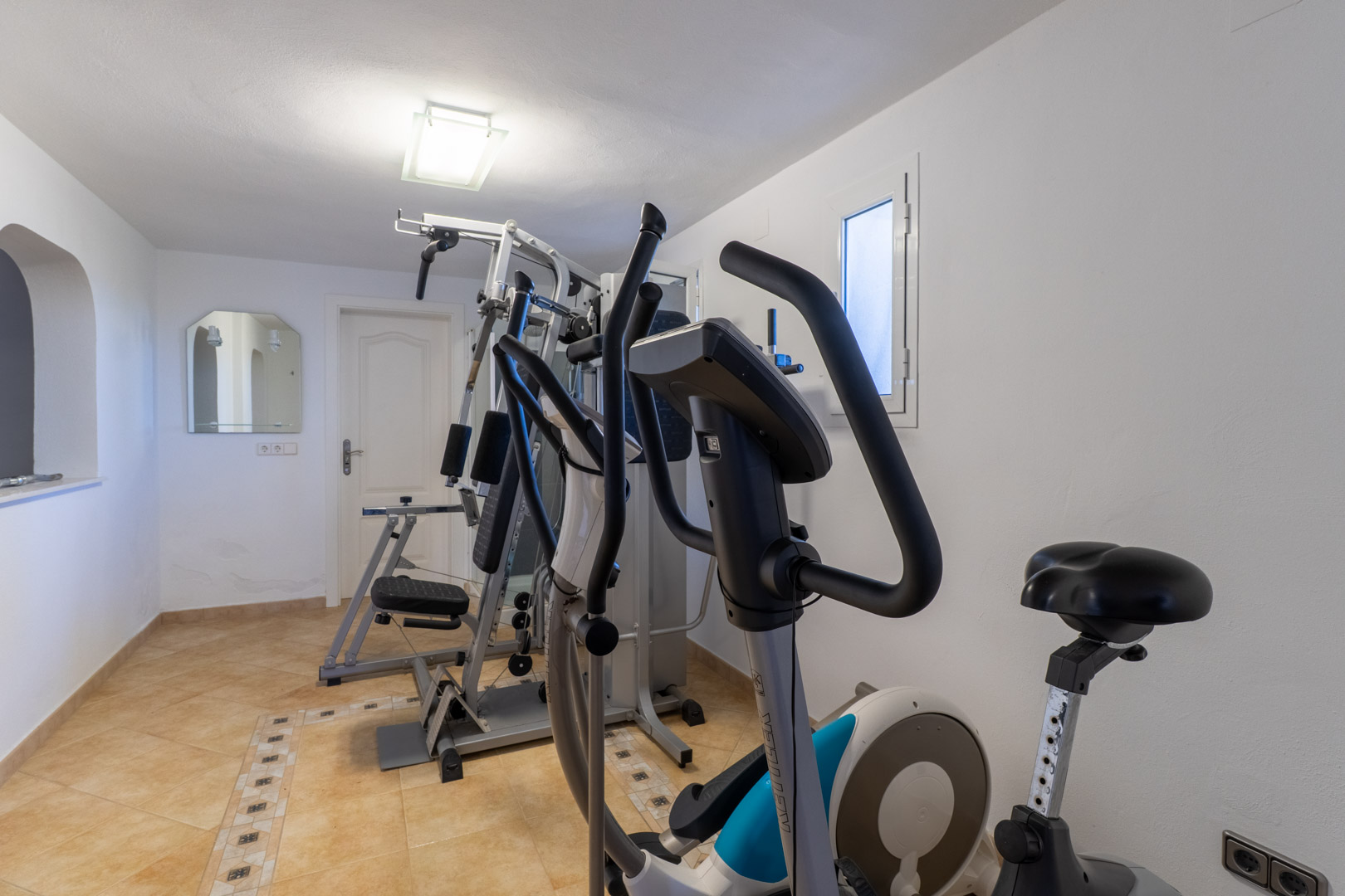 Privé fitnessruimte uitgerust met een krachtstation, crosstrainer en hometrainer op een betegelde vloer met sierrand.