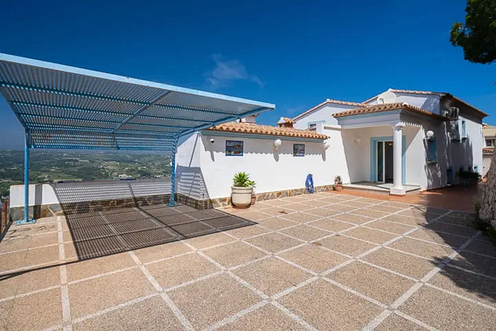 Ruime buitenparkeerplaats met tegelvloer, een blauwe metalen carport en vrij uitzicht over de omliggende bergen en het mediterrane landschap.