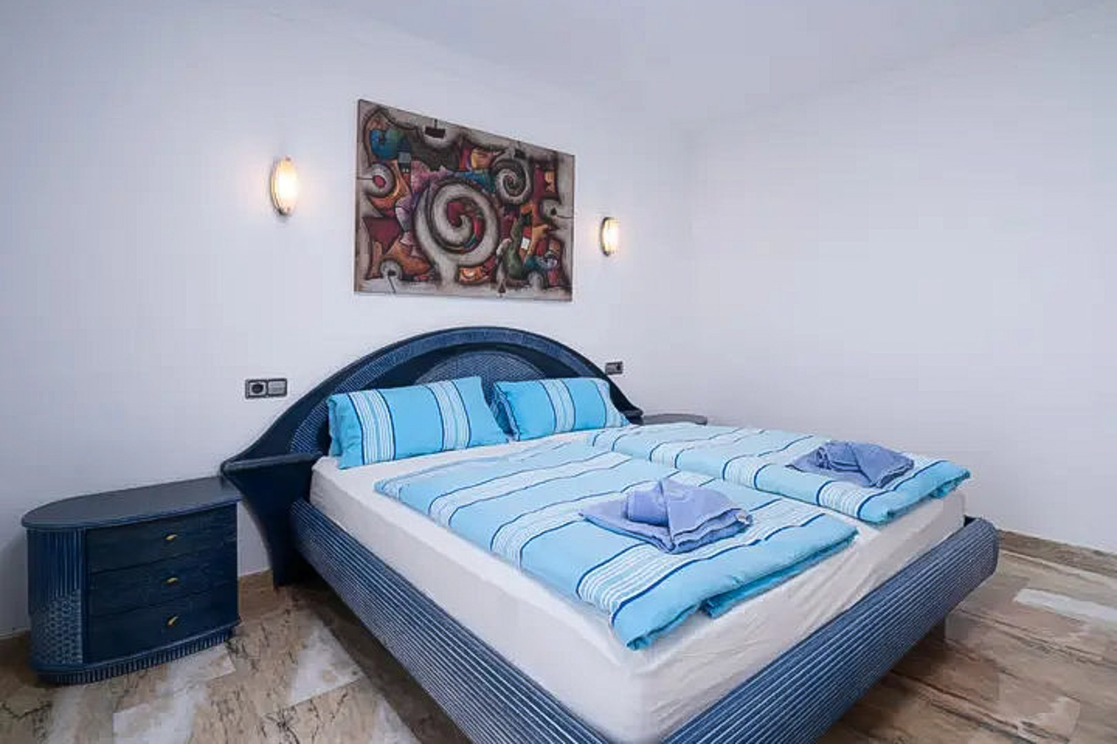 Slaapkamer met een blauw rieten hoofdbord en bijpassend nachtkastje. Voorzien van een gepolijste marmeren vloer, wandlampen en abstracte wanddecoratie.