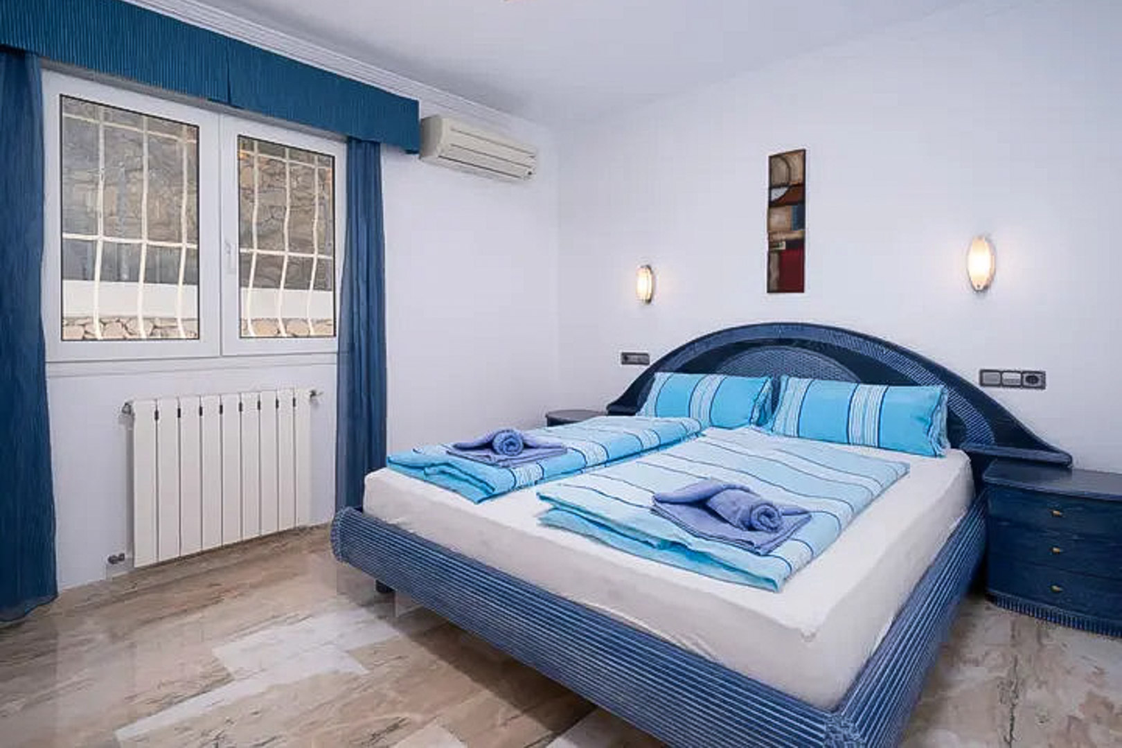 Slaapkamer met gepolijste marmeren vloer, tweepersoonsbed in blauwtinten, airconditioning, radiator en een beveiligd raam met bijpassende gordijnen.