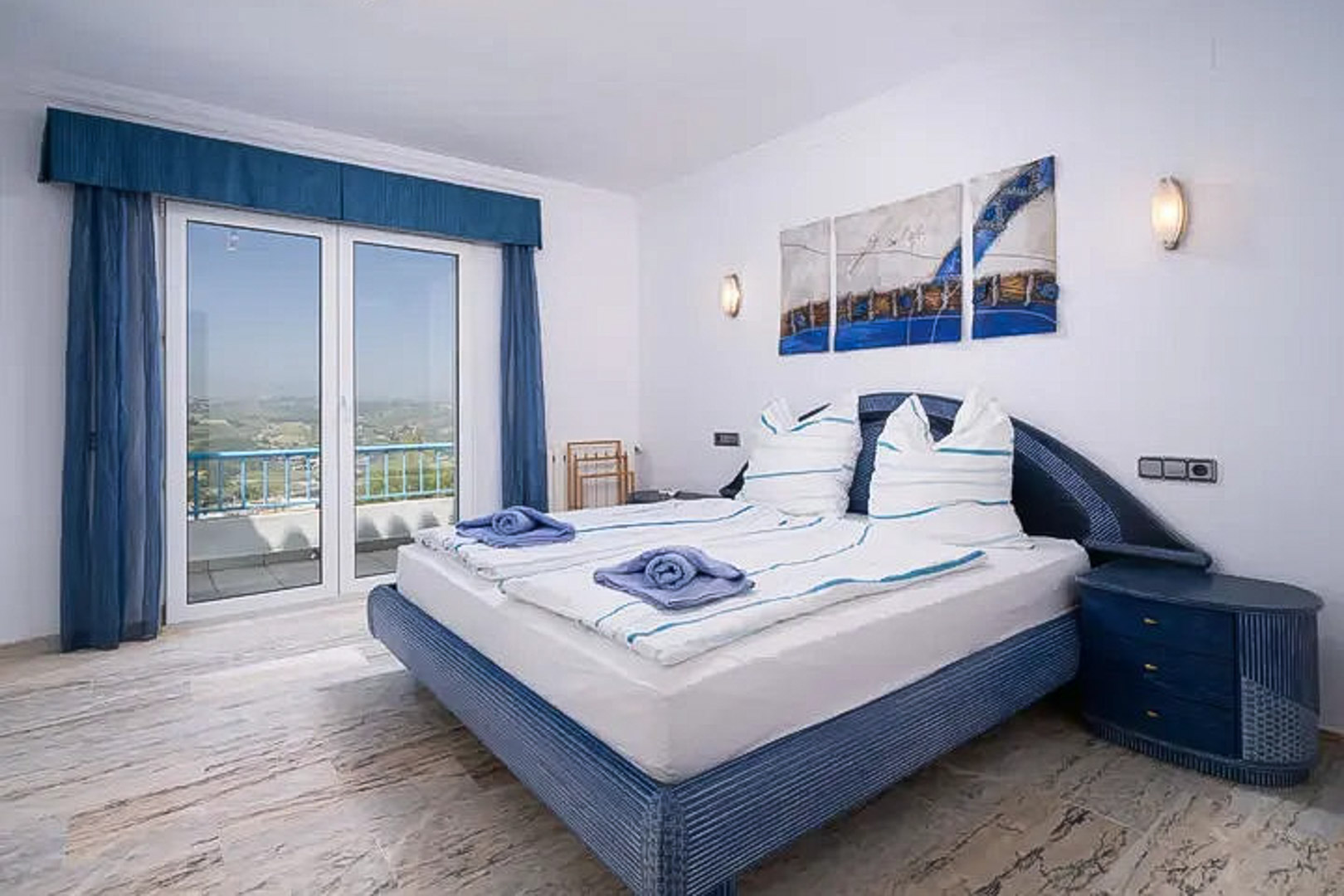 Slaapkamer met marmeren vloer, tweepersoonsbed in blauwtinten en directe toegang tot een balkon met panoramisch uitzicht over de omgeving.