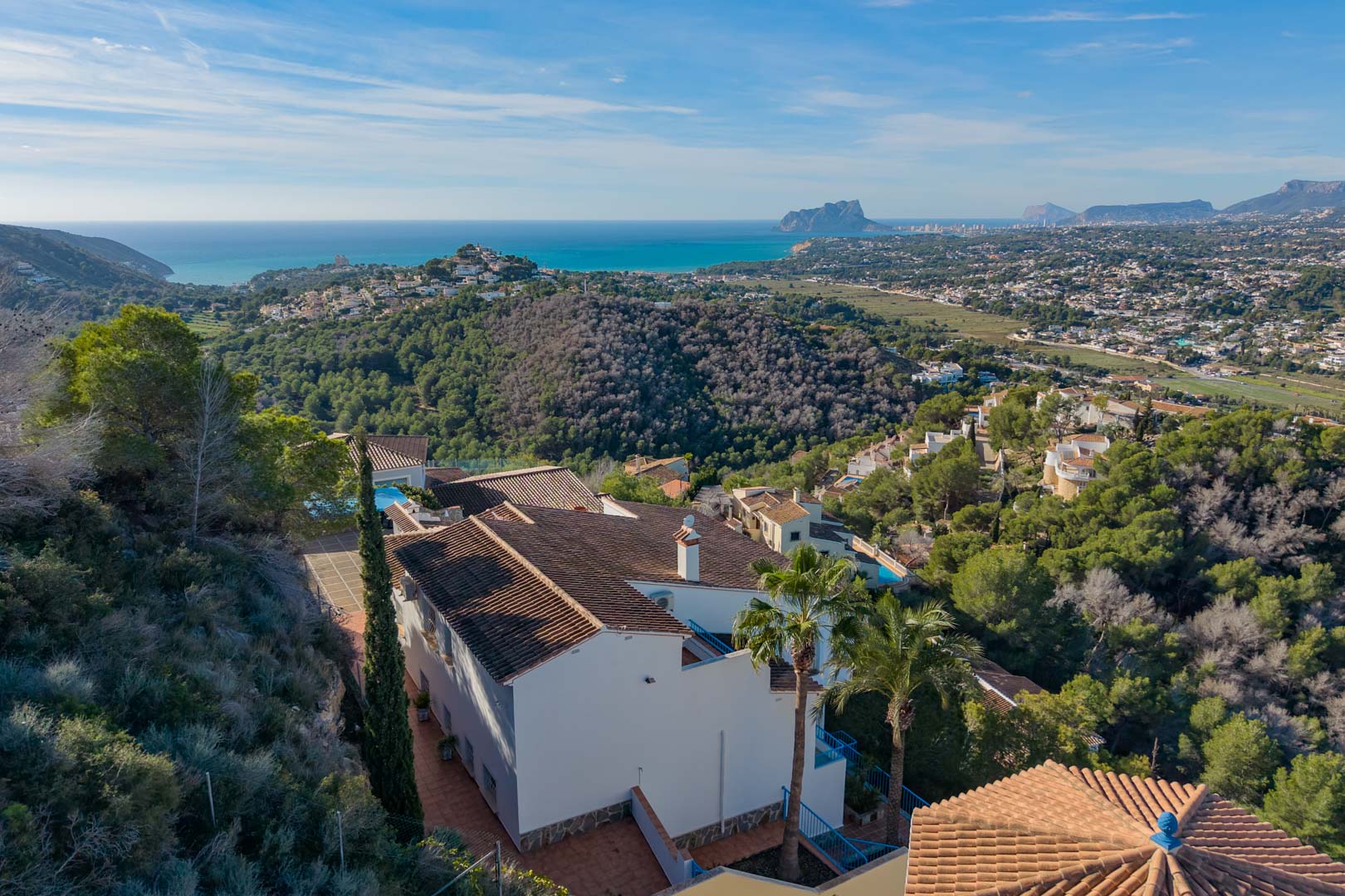 Verhoogd zicht op de villa met mediterrane architectuur, omgeven door pijnbomen met een panoramisch uitzicht over de kustlijn en de Peñón de Ifach.