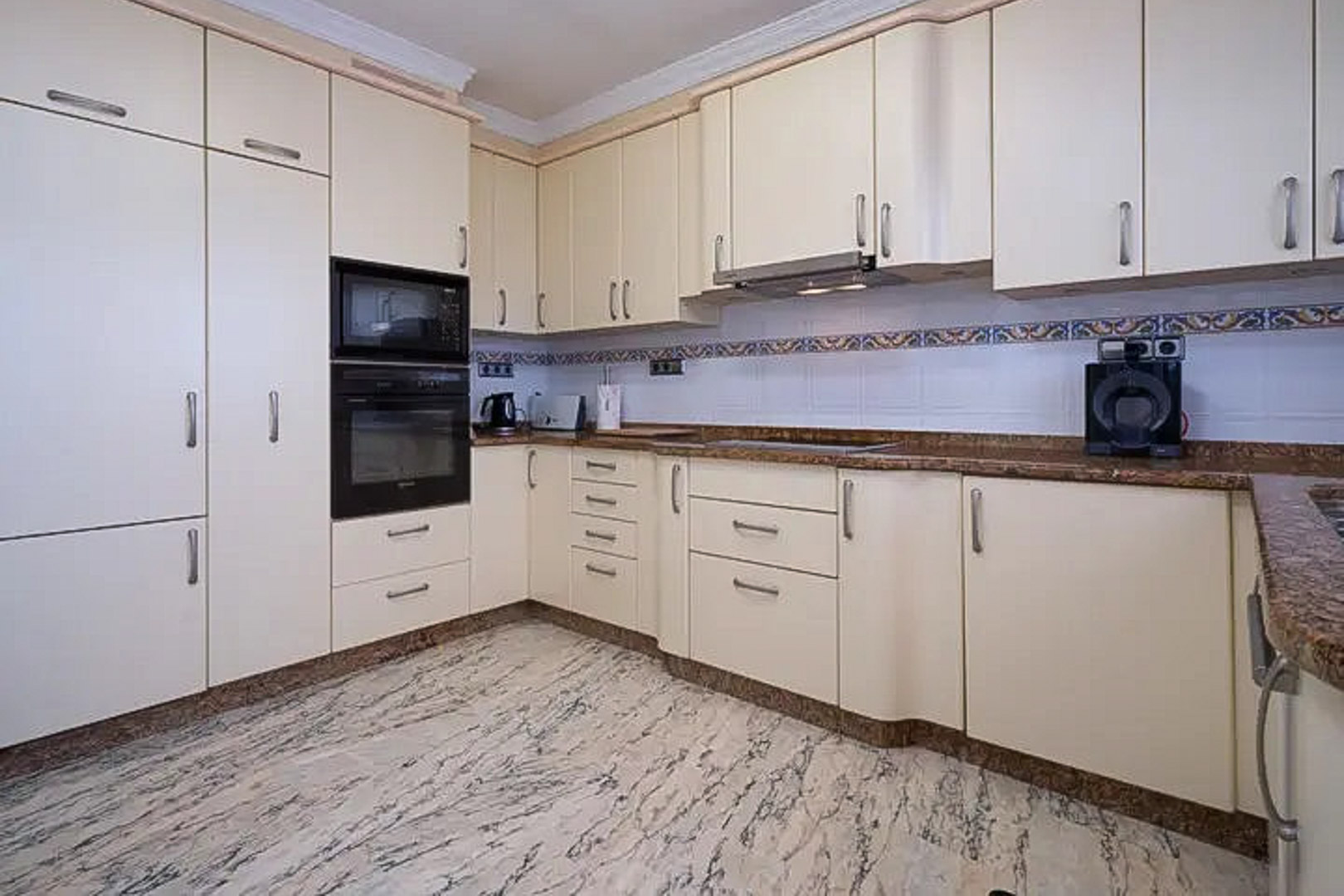 Volledig uitgeruste keuken met crèmekleurige kasten, bruin granieten werkbladen en inbouwapparatuur. Afgewerkt met een geaderde marmeren vloer en een decoratieve tegelwand.