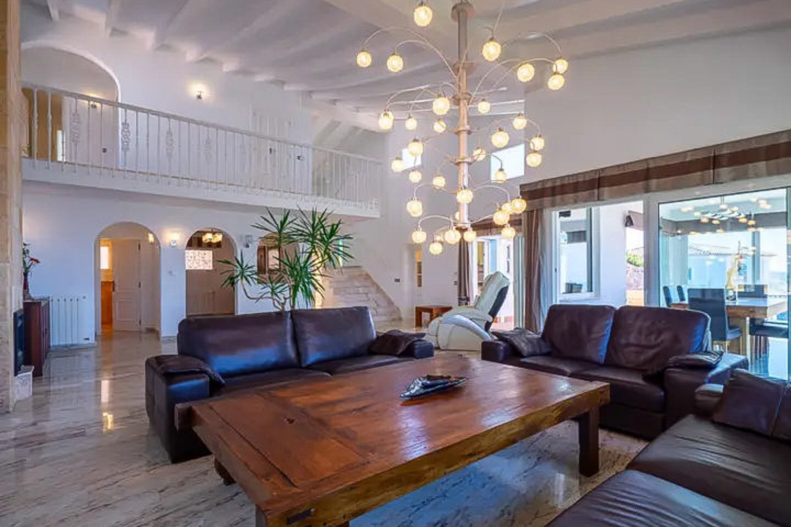Woonkamer met dubbele plafondhoogte, donkerlederen banken, een massief houten salontafel en designkroonluchter. Marmeren vloeren en een witte mezzanine sieren de ruimte.