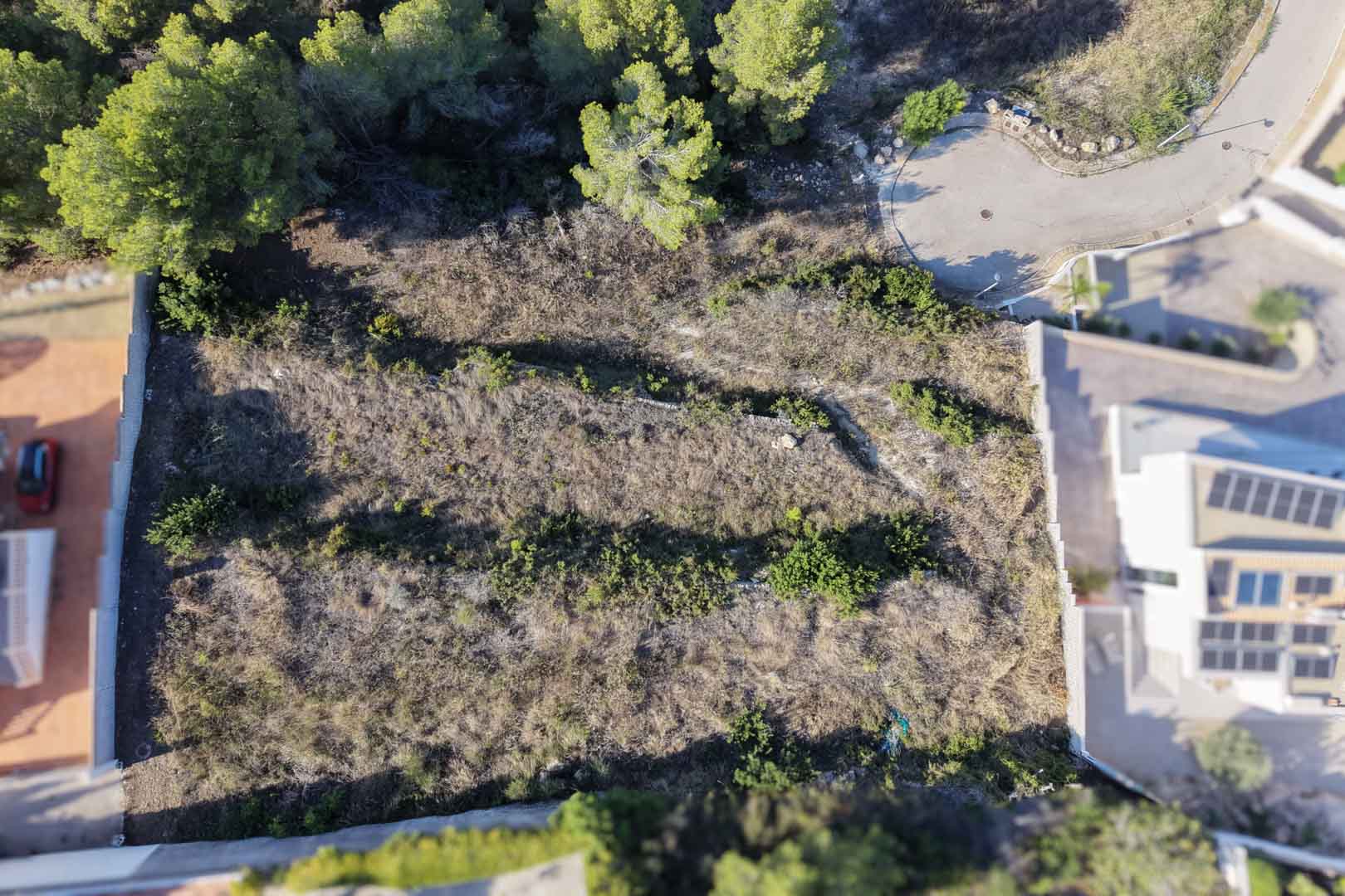 Bovenaanzicht van het terrasvormige bouwperceel met natuurstenen muren, omringd door pijnbomen en gelegen aan een rustige doodlopende straat.
