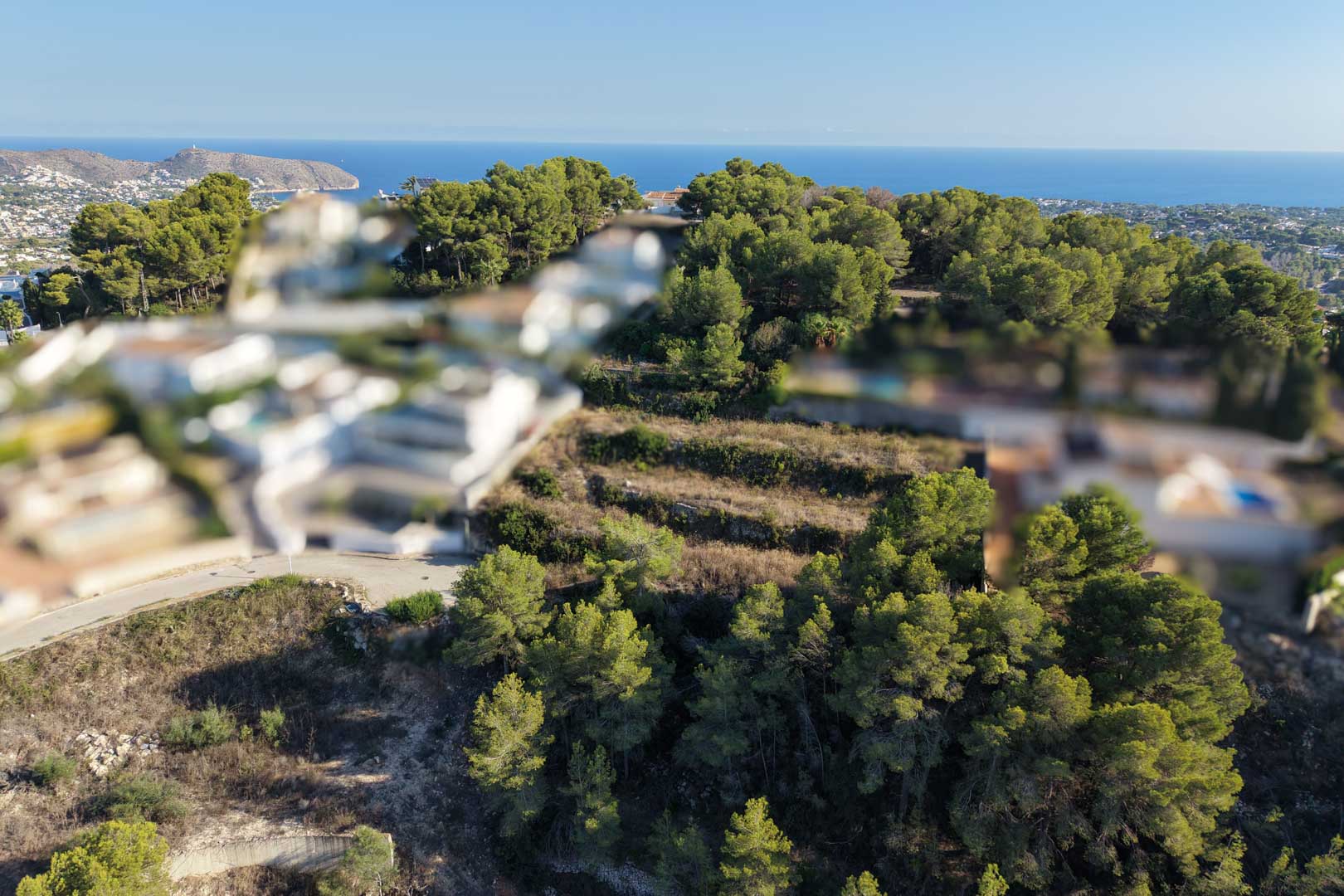 Luchtfoto van het terrasvormige bouwperceel, omringd door dichte mediterrane pijnbomen met vrij uitzicht op de zee en de kustlijn van Moraira.