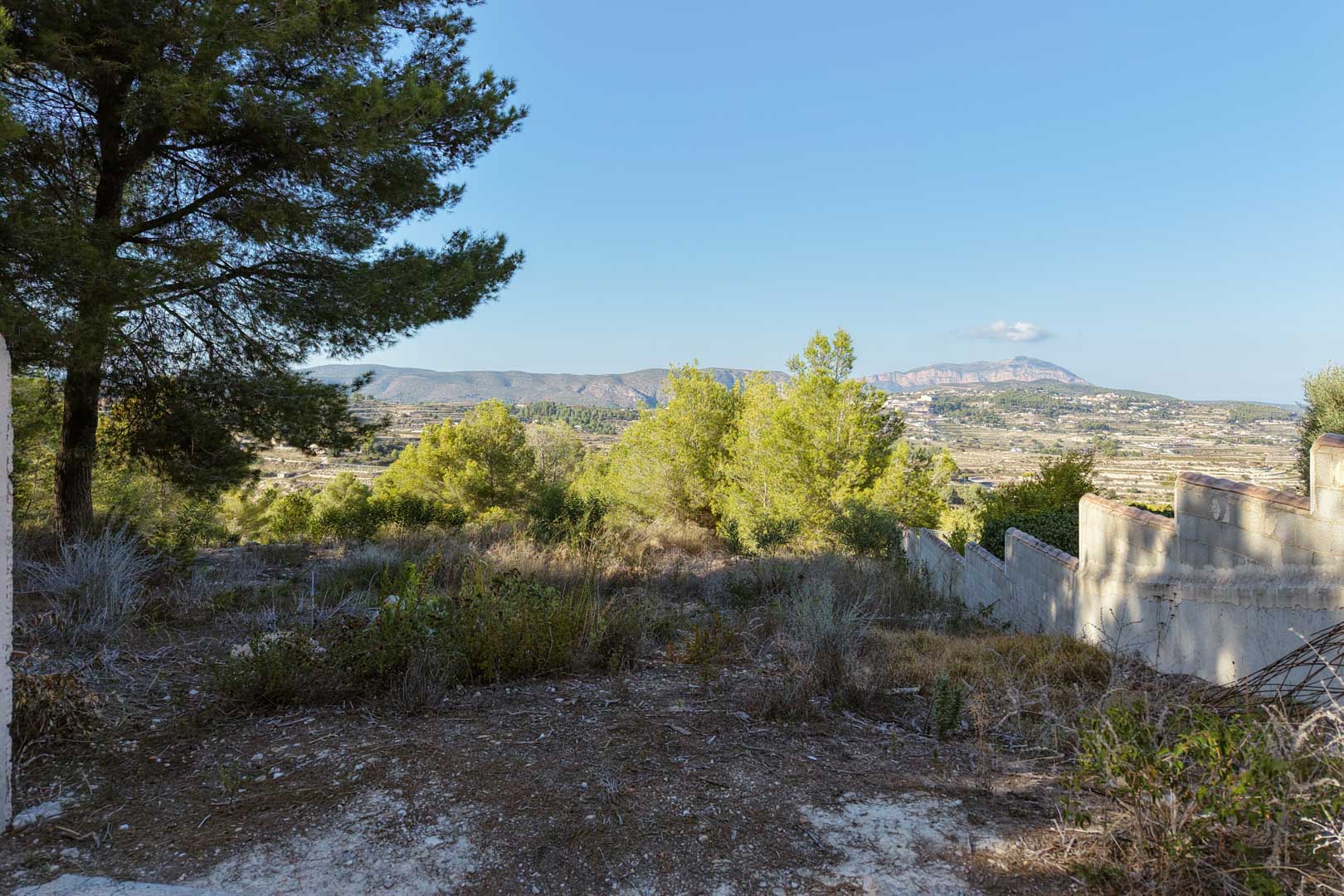 Hooggelegen perceel met panoramisch uitzicht op de vallei en bergen. Het terrein is voorzien van natuurlijke vegetatie, pijnbomen en een trapsgewijze omheiningsmuur.