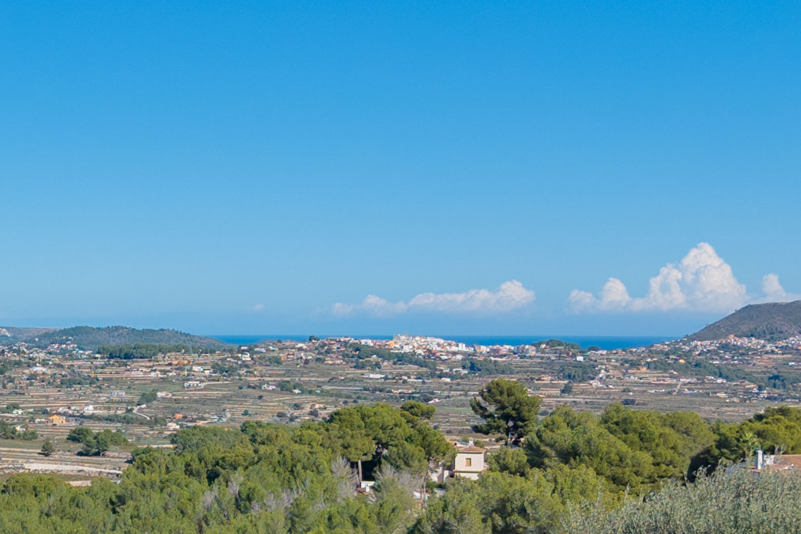 Panoramisch uitzicht vanaf het perceel in Benimeit, met terrasvormige heuvels, mediterrane dennenbossen en de verre zee onder een helderblauwe lucht.