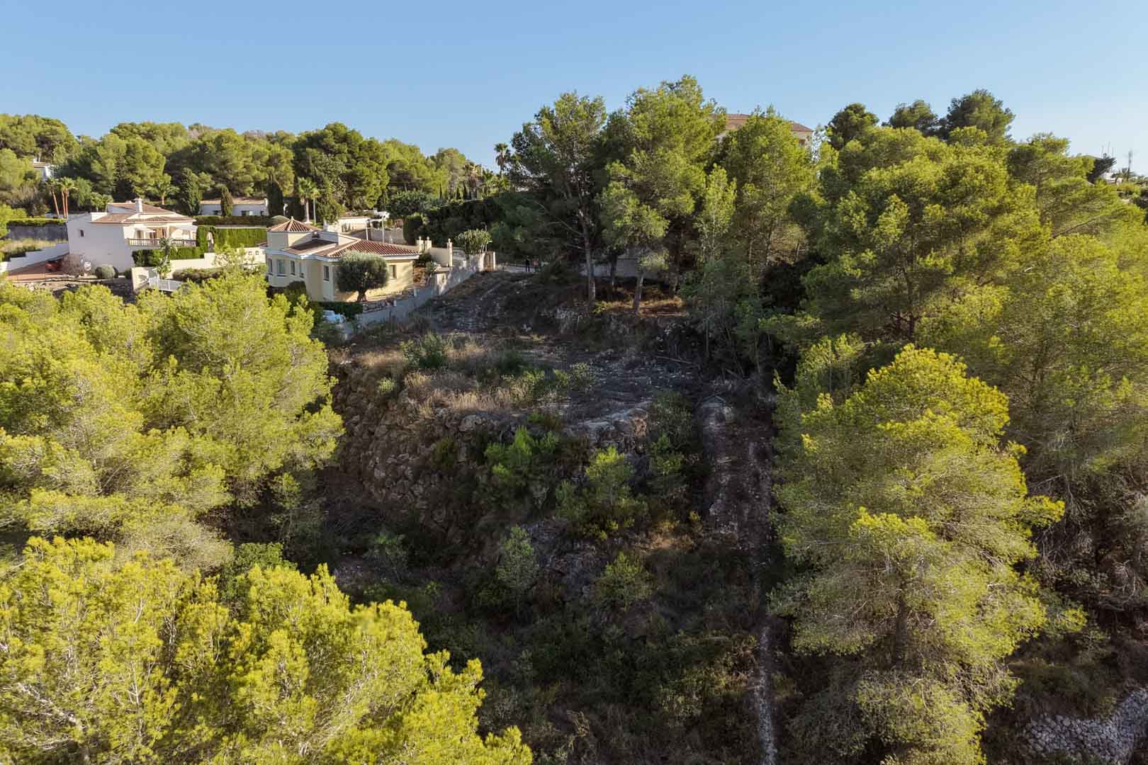 Verhoogd bouwperceel in Benimeit, omgeven door volgroeide pijnbomen en mediterrane vegetatie, met natuurlijk terrasvormig terrein en uitzicht op naburige villa's.