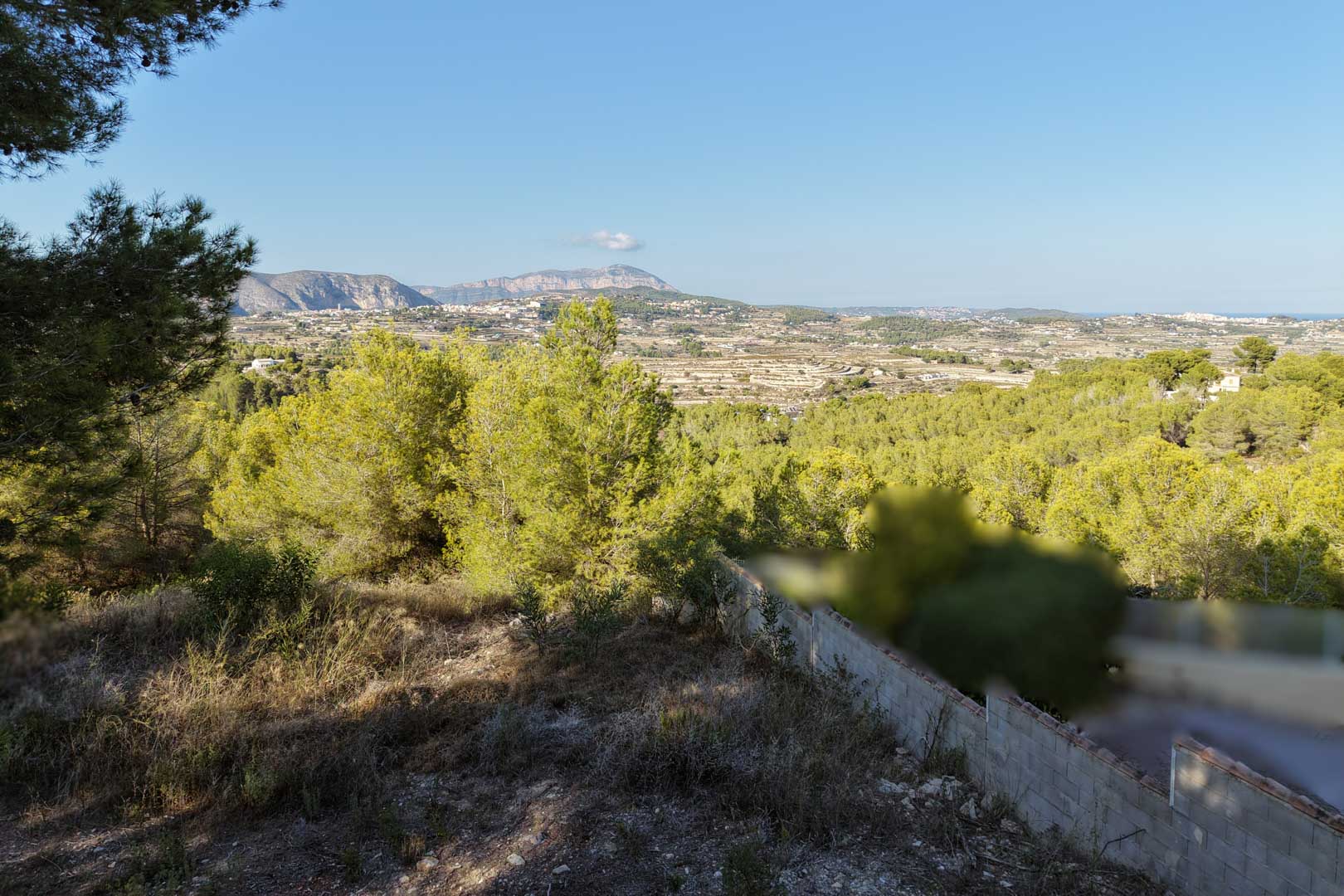 Verhoogd panoramisch uitzicht vanaf het perceel over de vallei van Moraira, met inheemse dennenbossen en verre bergketens onder een heldere blauwe hemel.