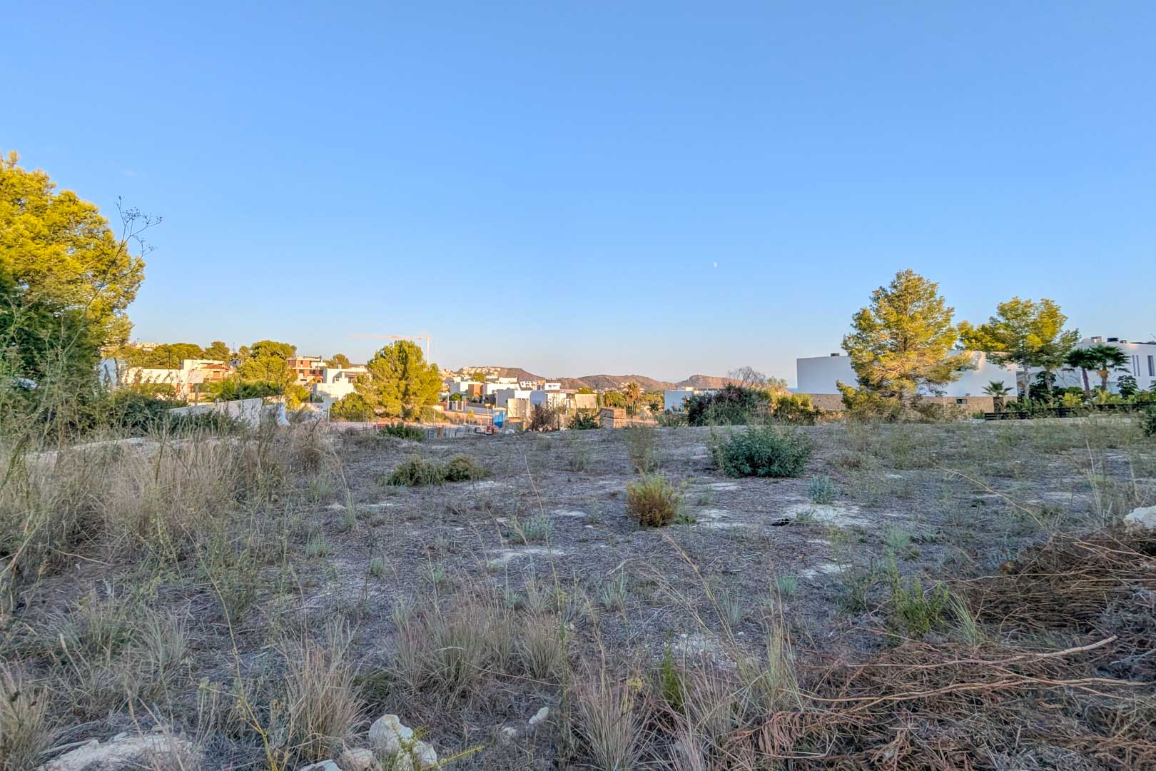 Panoramisch uitzicht op het vlakke bouwperceel in El Tesoro, omgeven door mediterrane begroeiing met zicht op moderne witte villa's en heuvels.