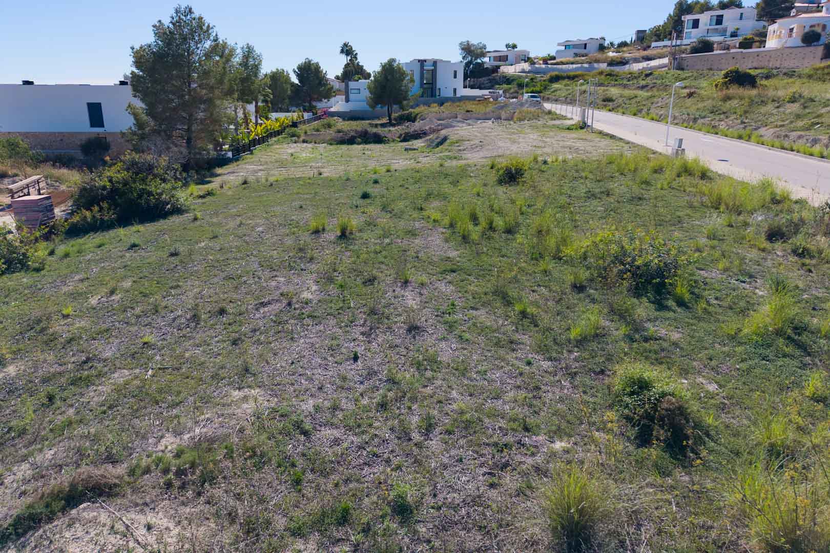 Vlak stedelijk perceel in El Tesoro, Moraira, met geasfalteerde toegangsweg en uitzicht op moderne villa's. Het terrein biedt een vrij uitzicht over de vallei.