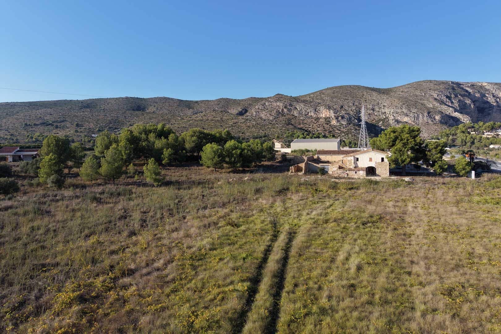 Luchtfoto van het traditionele stenen huis en het uitgestrekte vlakke perceel, omgeven door mediterrane vegetatie met de bergen van Teulada op de achtergrond.