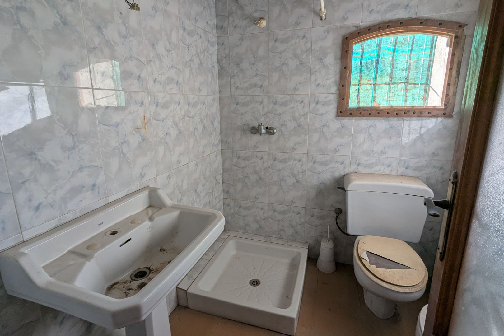 Originele badkamer met blauwe marmerlook wandtegels, een wastafel op zuil, vierkante douchebak en een klein boogvenster met houten kozijn.