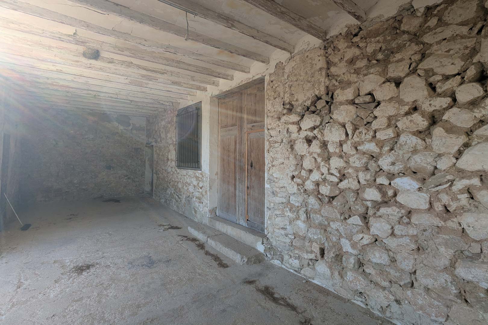 Overdekte veranda met muren van natuursteen, originele houten plafondbalken en een authentieke dubbele houten toegangsdeur in traditionele stijl.