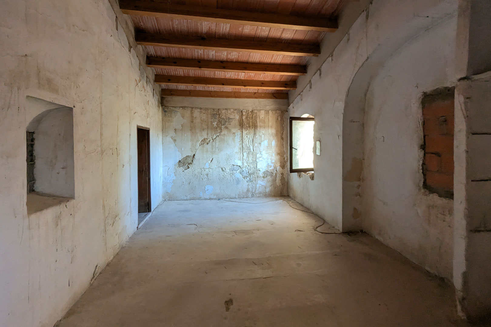 Ruime open kamer met hoge plafonds voorzien van houten balken en terracotta tegels. De dikke muren tonen originele nissen en een zijraam dat natuurlijk licht binnenlaat.