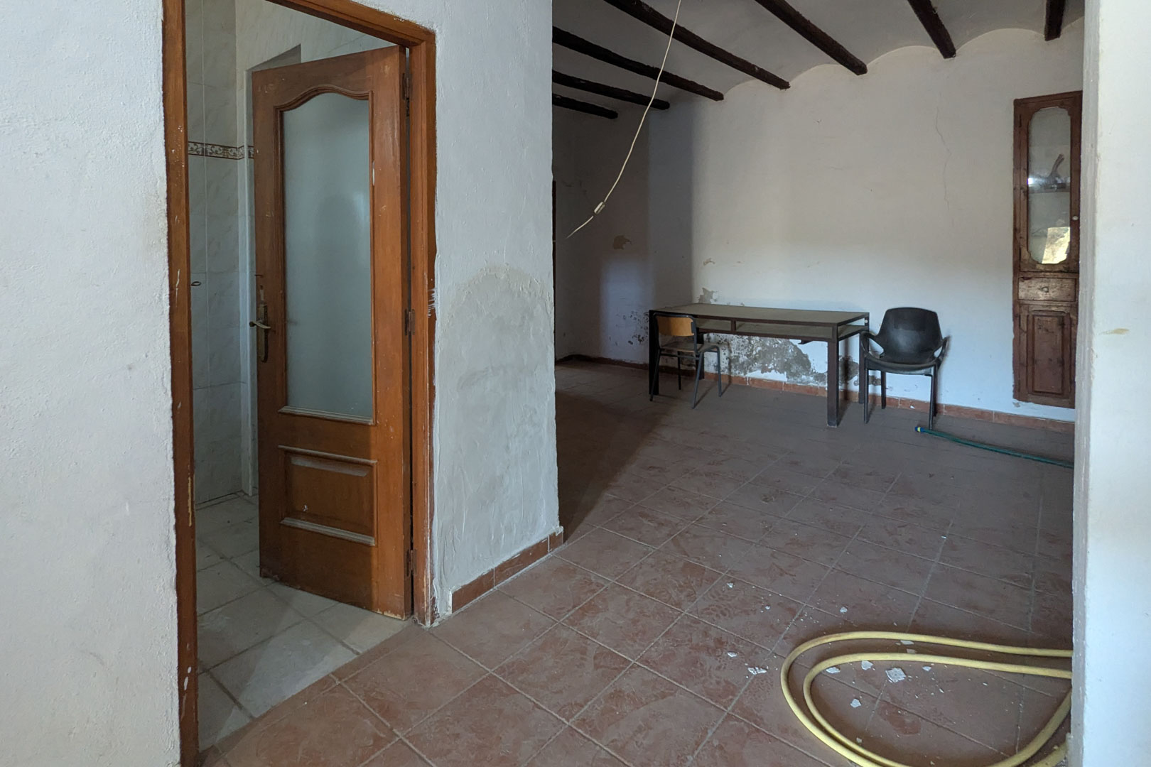 Rustiek interieur met donkere houten plafondbalken, terracotta vloertegels en witgekalkte muren. Voorzien van een houten deur met glas en een ingebouwde muurkast.