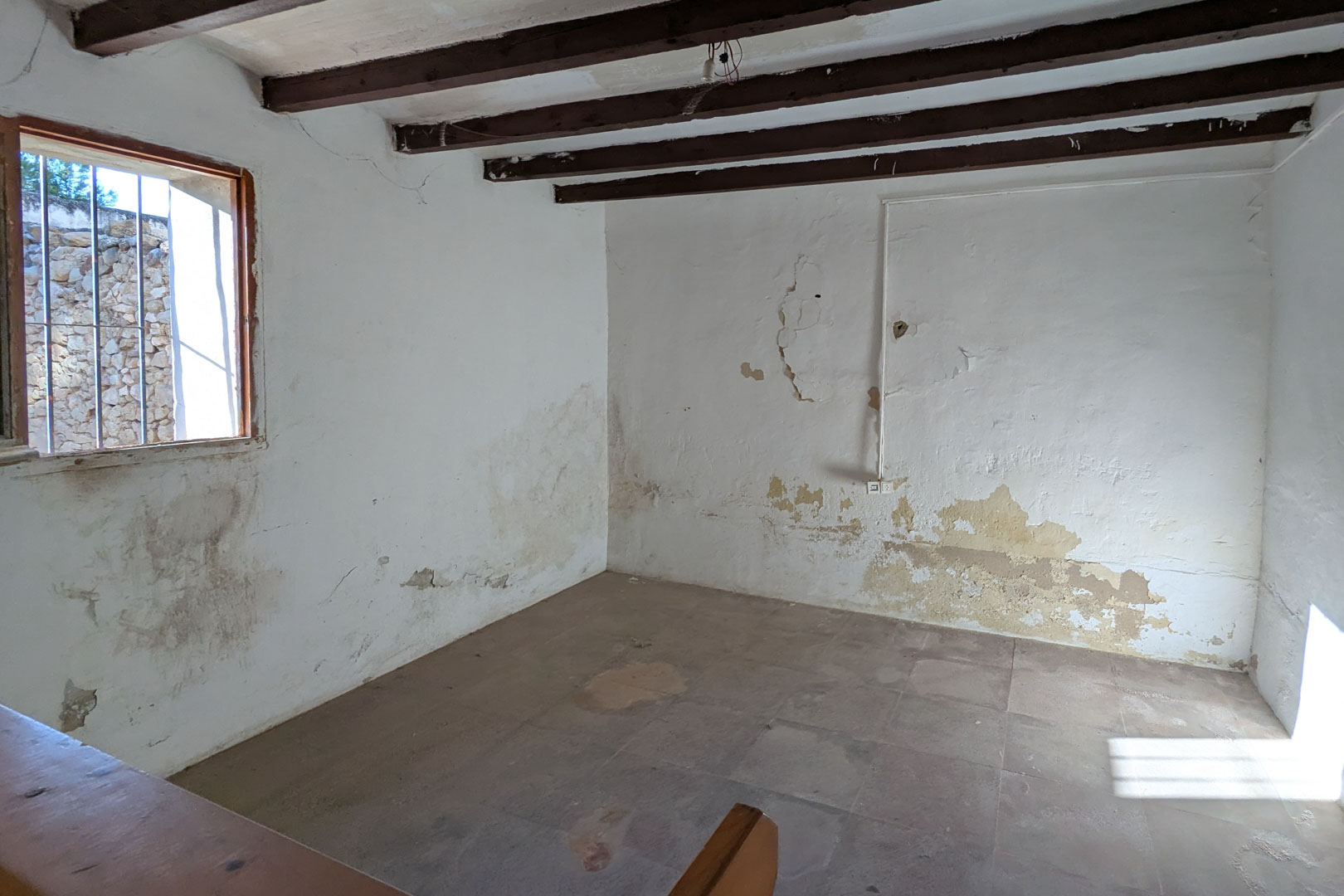 Rustieke kamer met originele donkere houten balken, witgekalkte muren met authentieke gebruikssporen en een tegelvloer. Een raam met tralies zorgt voor natuurlijk licht en zicht op een stenen muur.
