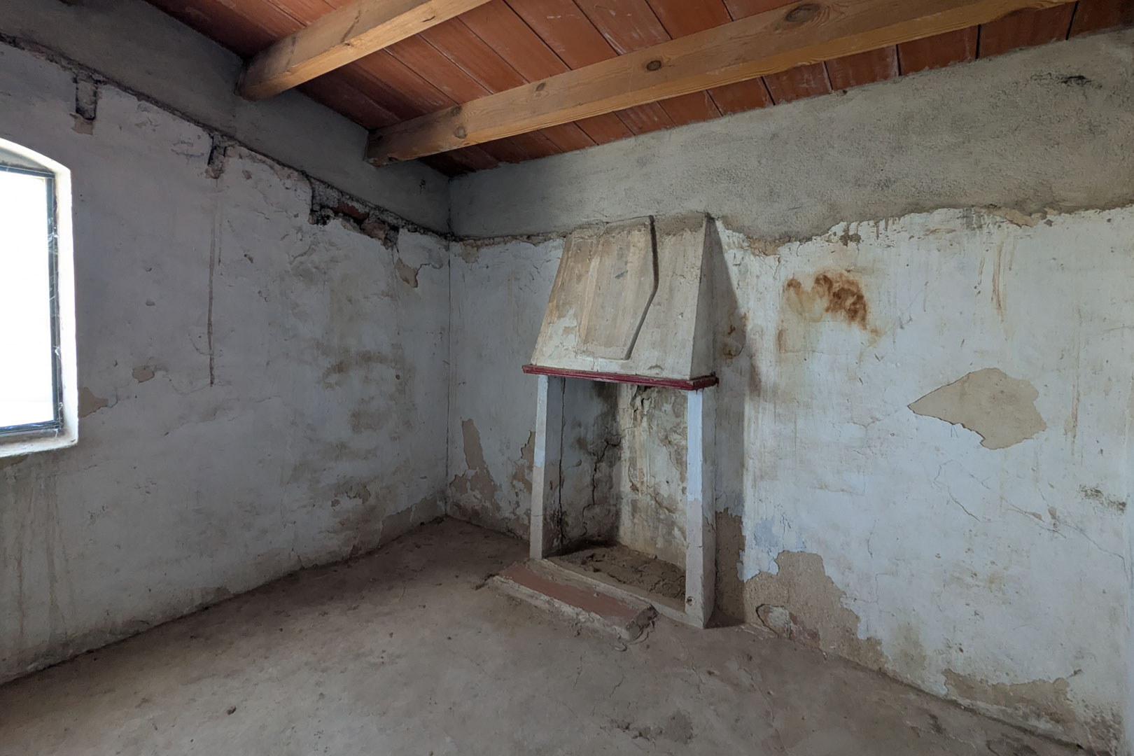 Rustieke woonkamer met een traditionele schouw, zichtbare houten plafondbalken en authentieke muren met kalkpleister die wachten op renovatie.