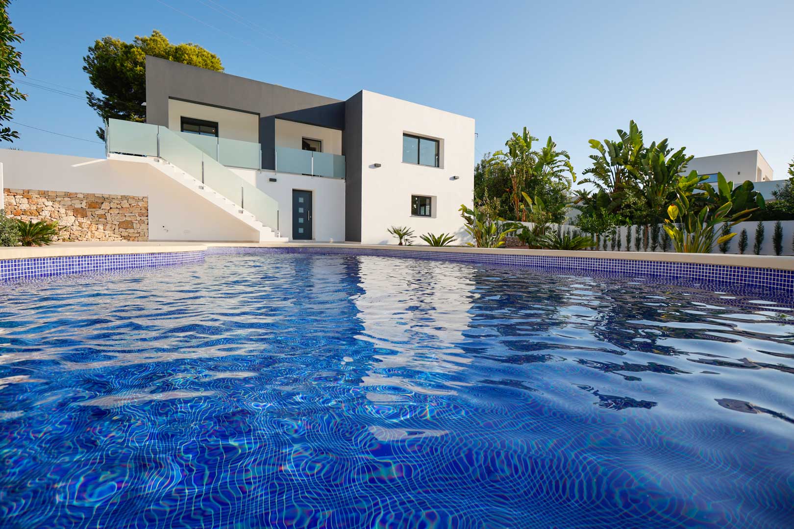 Buitenaanzicht van de villa met blauw betegeld zwembad, minimalistische witte en grijze gevel, natuurstenen muren en terrassen met glazen balustrades.