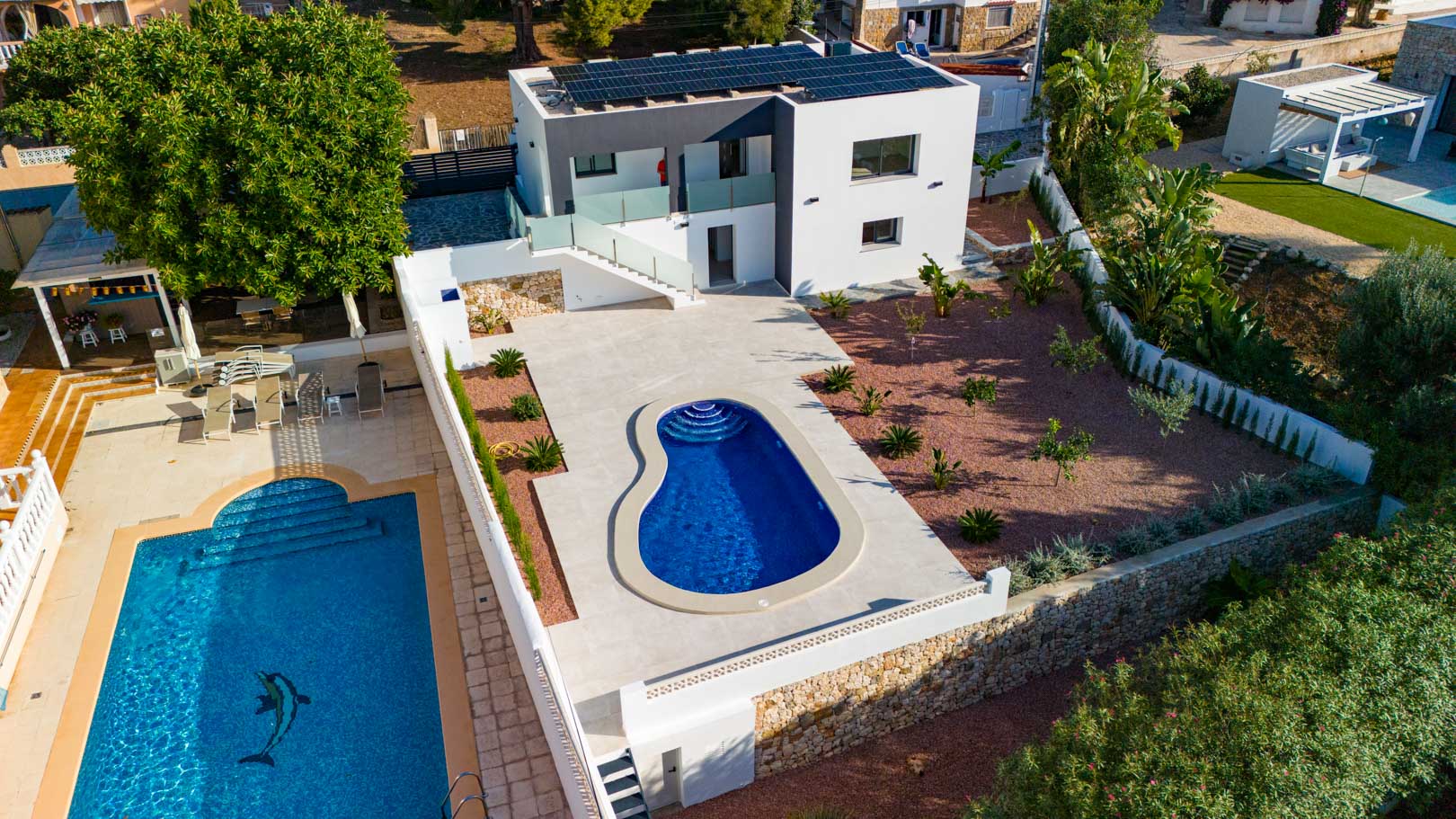 Luchtfoto van de moderne villa met zonnepanelen, organisch gevormd zwembad, rode grindtuin en natuurstenen muren. Glazen balustrades accentueren het eigentijdse ontwerp.