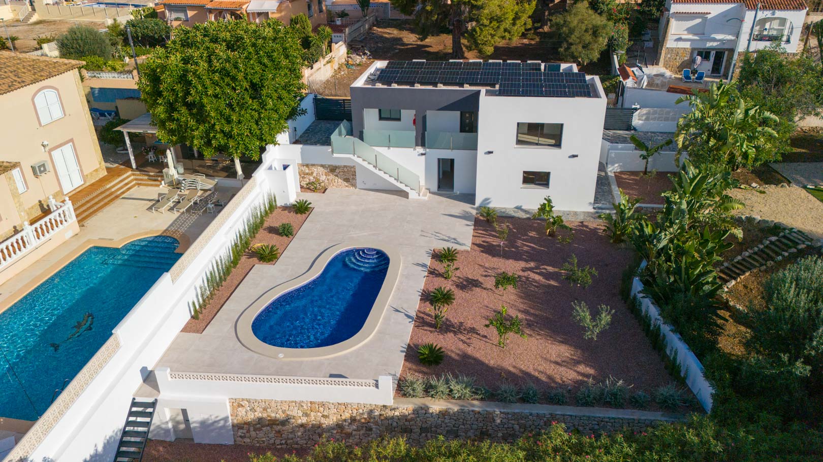 Luchtfoto van de villa met niervormig zwembad, rode grindtuin en zonnepanelen op het dak. De glazen balustrades en natuurstenen muren accentueren het moderne ontwerp.