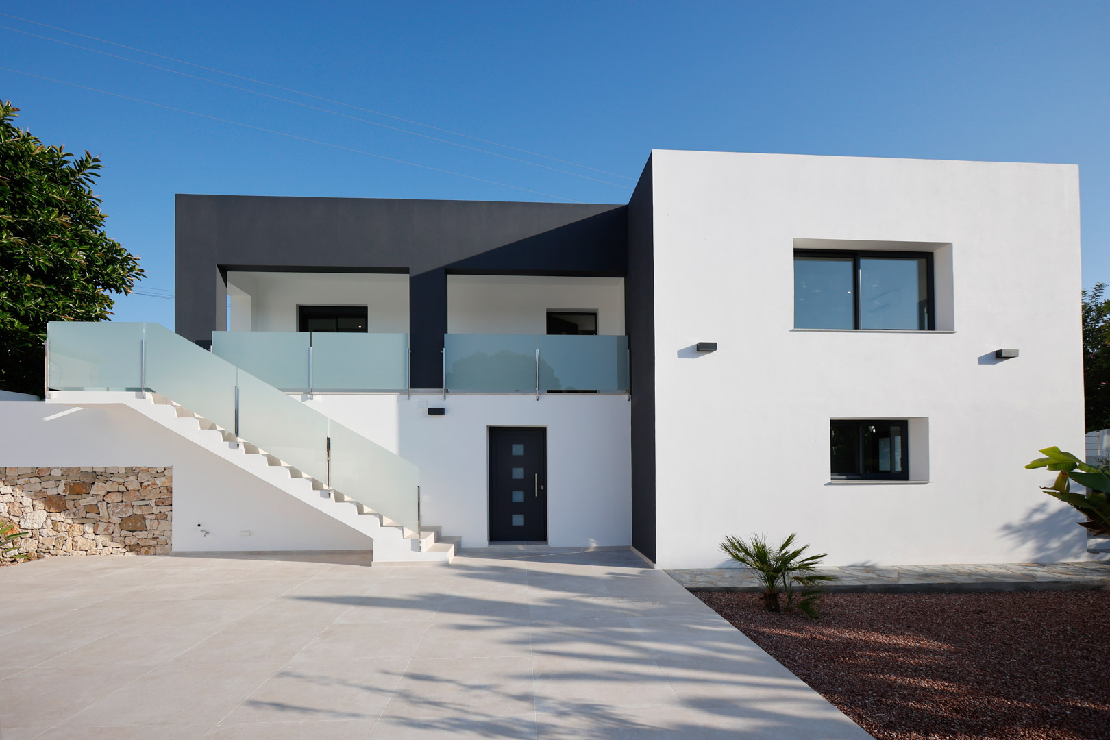 Minimalistische wit-grijze gevel met buitentrap, glazen balustrades, Technal aluminium kozijnen en een accentmuur van natuursteen.