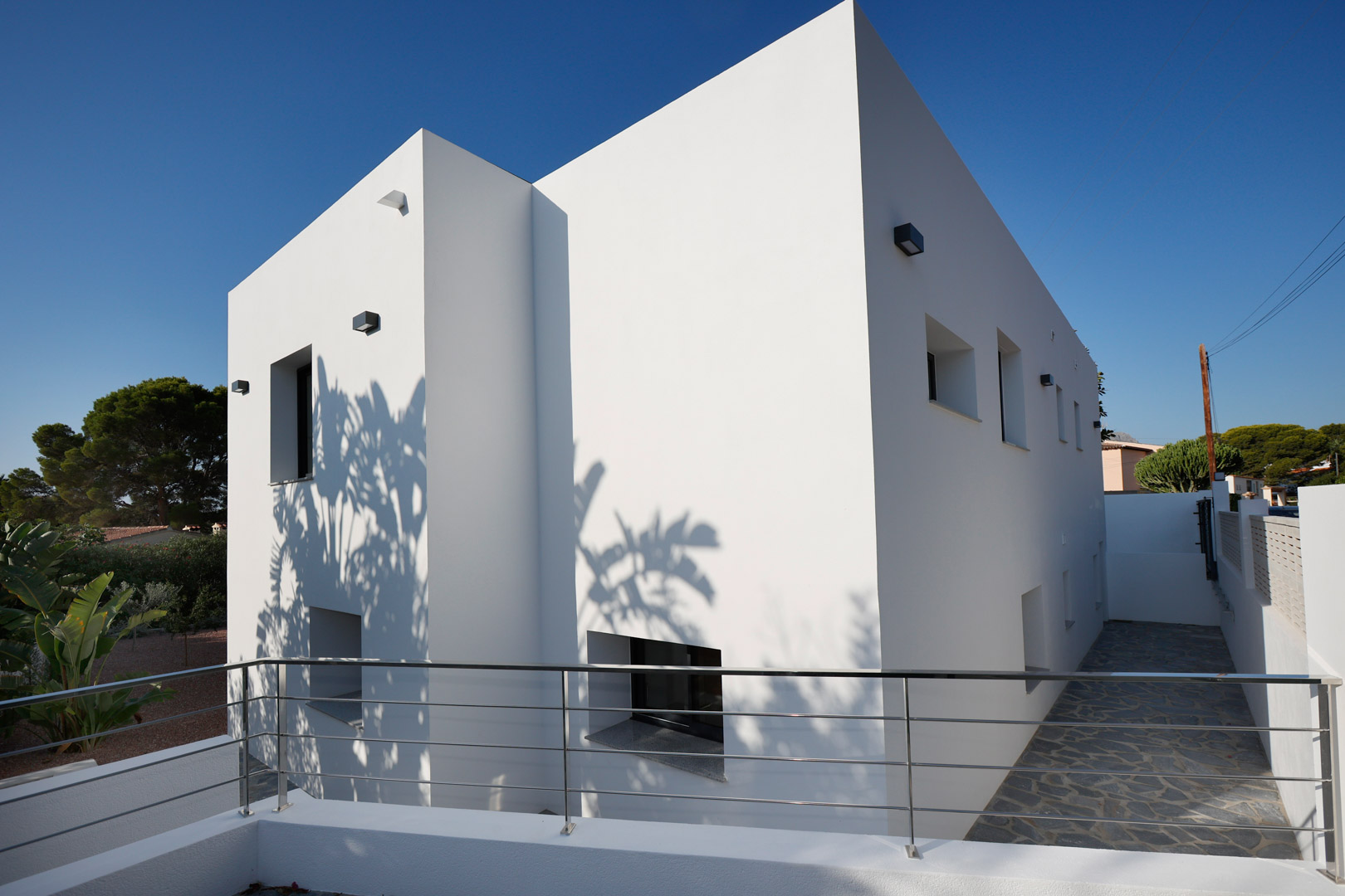 Minimalistische witte gevel met strakke geometrische lijnen, roestvrijstalen balustrades en natuurstenen bestrating. Schaduwen accentueren het eigentijdse ontwerp.