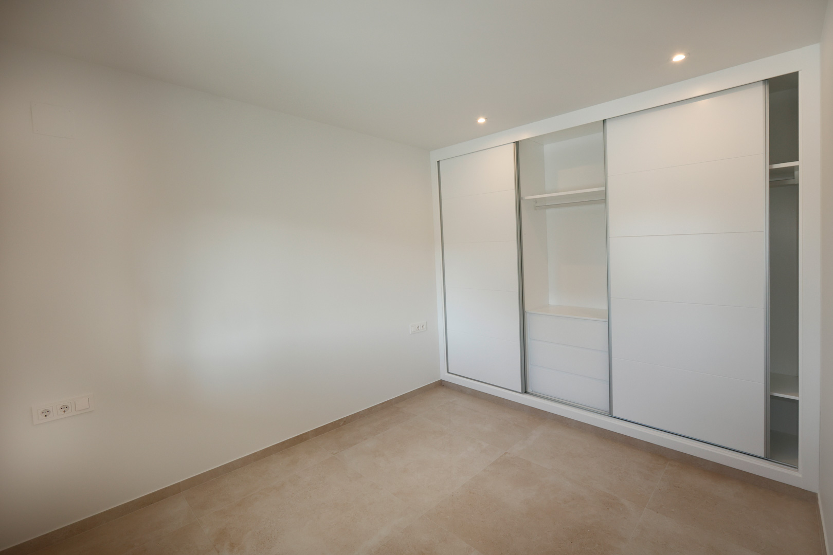 Slaapkamer met inbouwkast met wit gelakte schuifdeuren, porseleinen tegelvloer en inbouw-ledspots in het plafond.