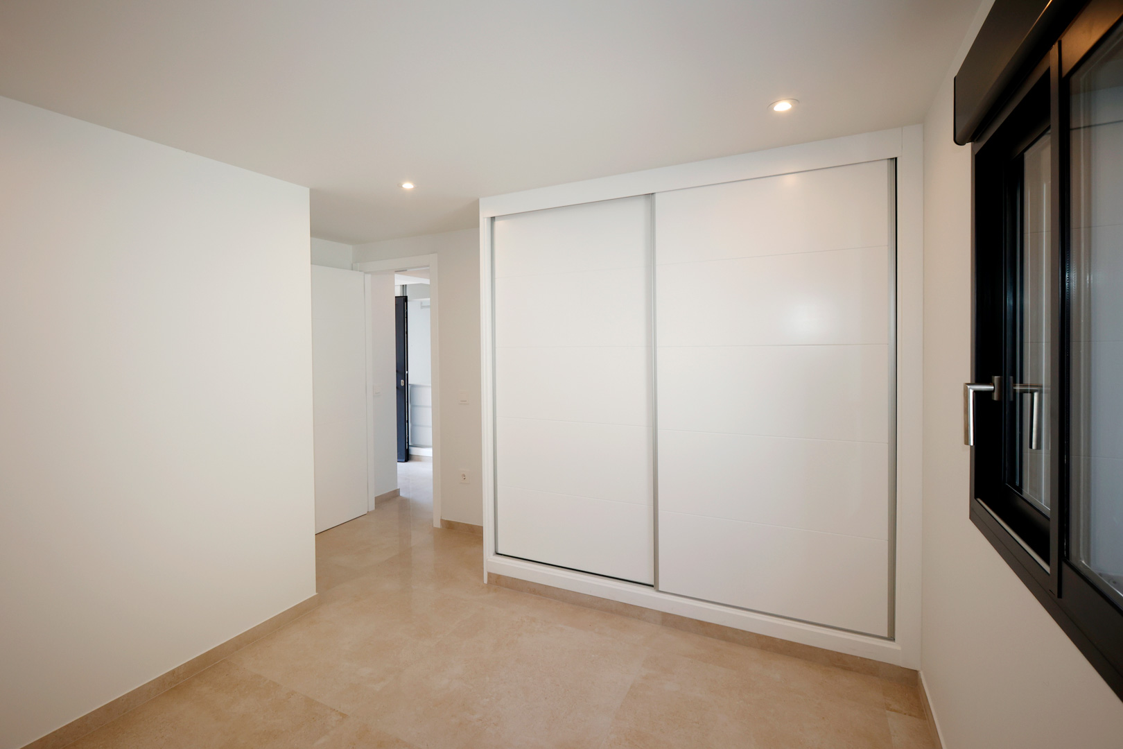 Slaapkamer met inbouwkast met witte schuifdeuren, porseleinen tegelvloer en inbouw-LED-verlichting naast een donker aluminium raamkozijn.
