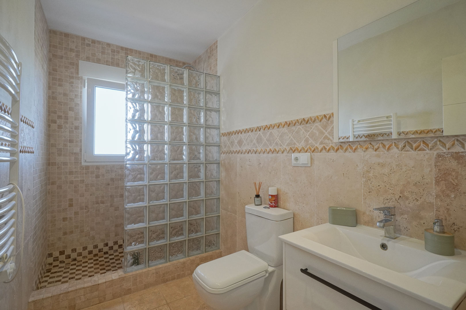 Badkamer met inloopdouche voorzien van glasblokken wand, aardetinten mozaïektegels, wit wastafelmeubel en handdoekradiator.