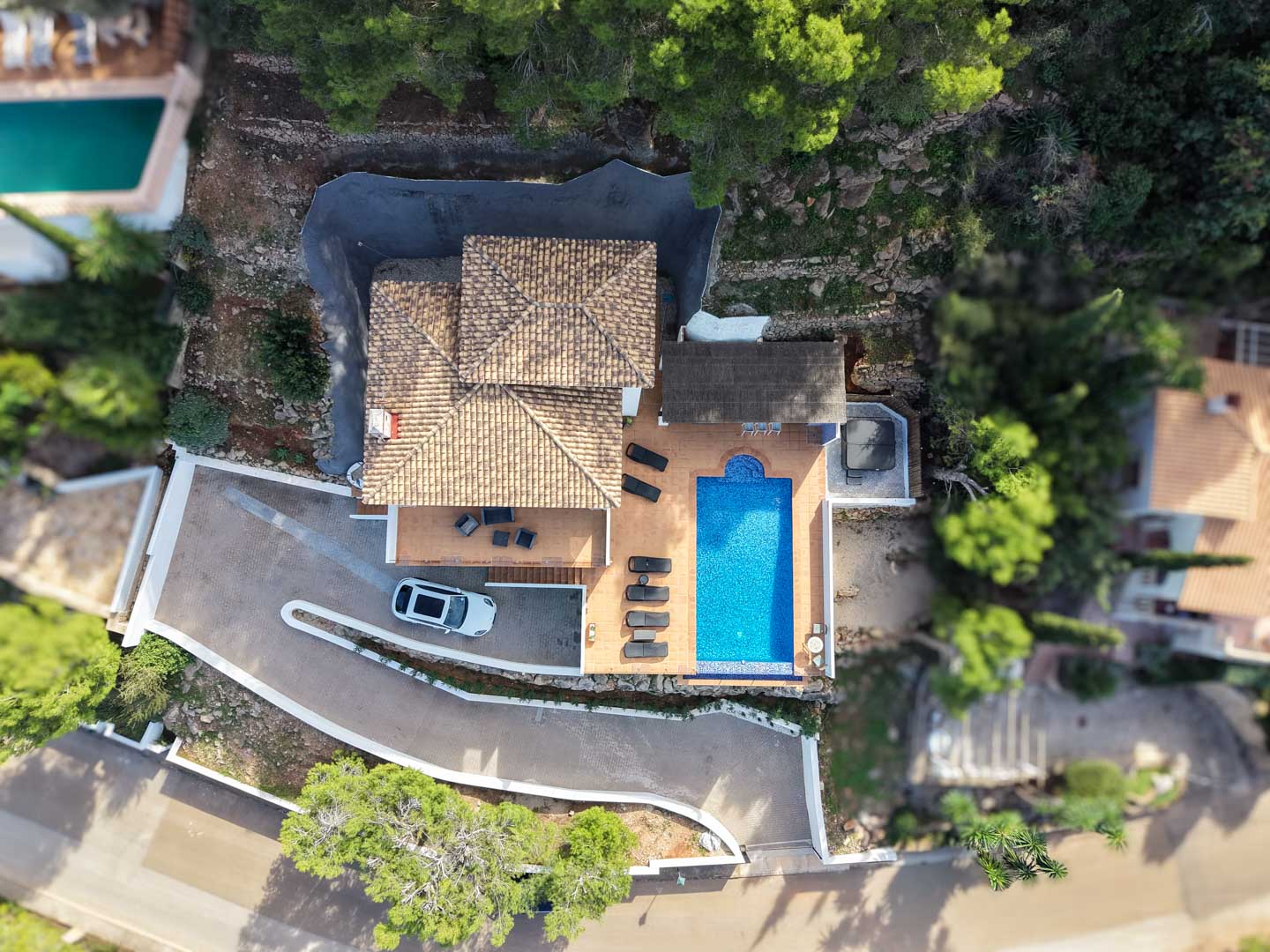 Bovenaanzicht van de villa met mediterraan pannendak, rechthoekig zwembad, zonneterras met ligstoelen, buitenjacuzzi en een eigen oprit omgeven door pijnbomen.