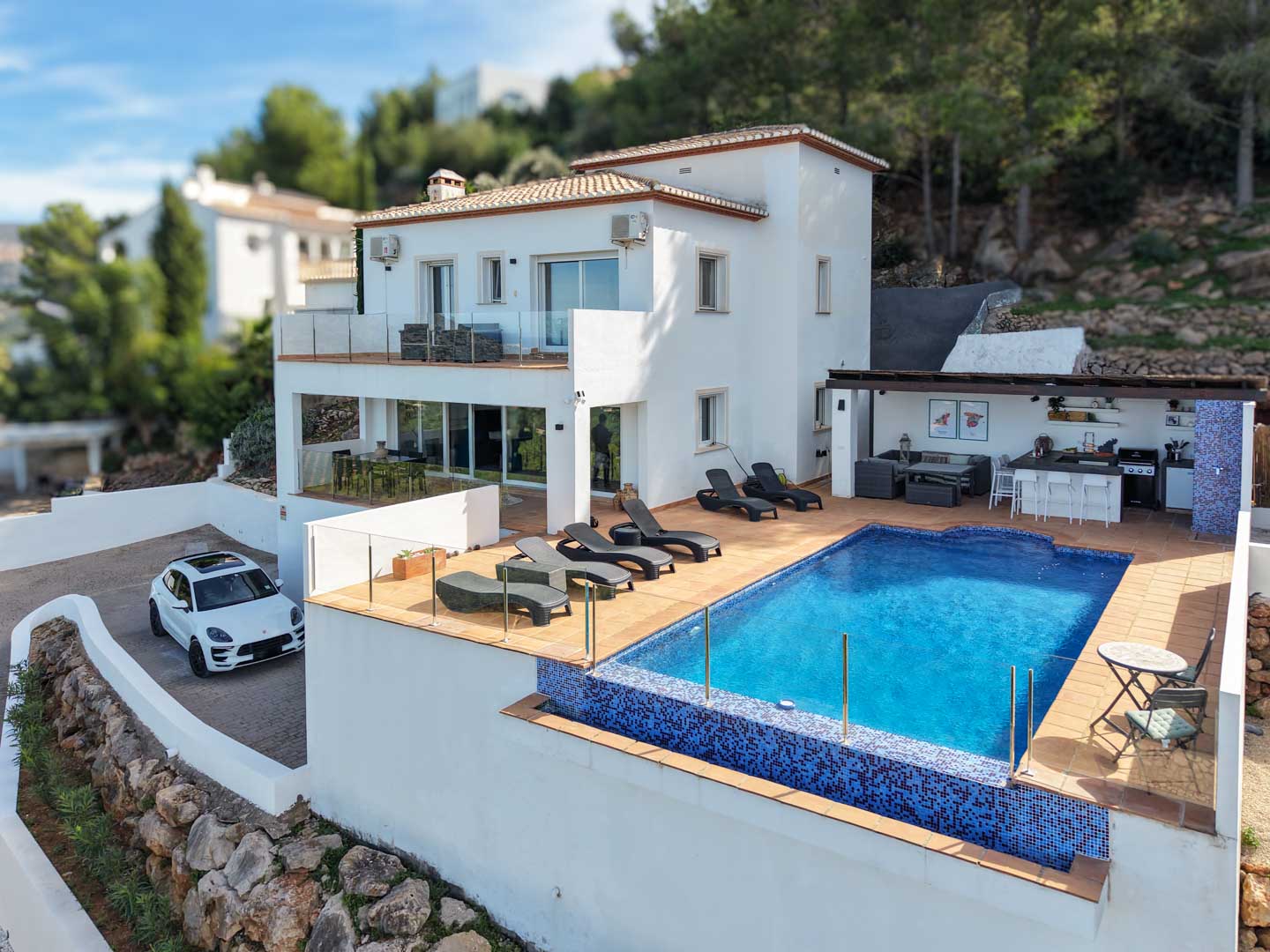 Buitenaanzicht van de villa met blauw mozaïekzwembad, zonneterras, buitenkeuken onder een pergola en witte mediterrane gevel op een verhoogd perceel.