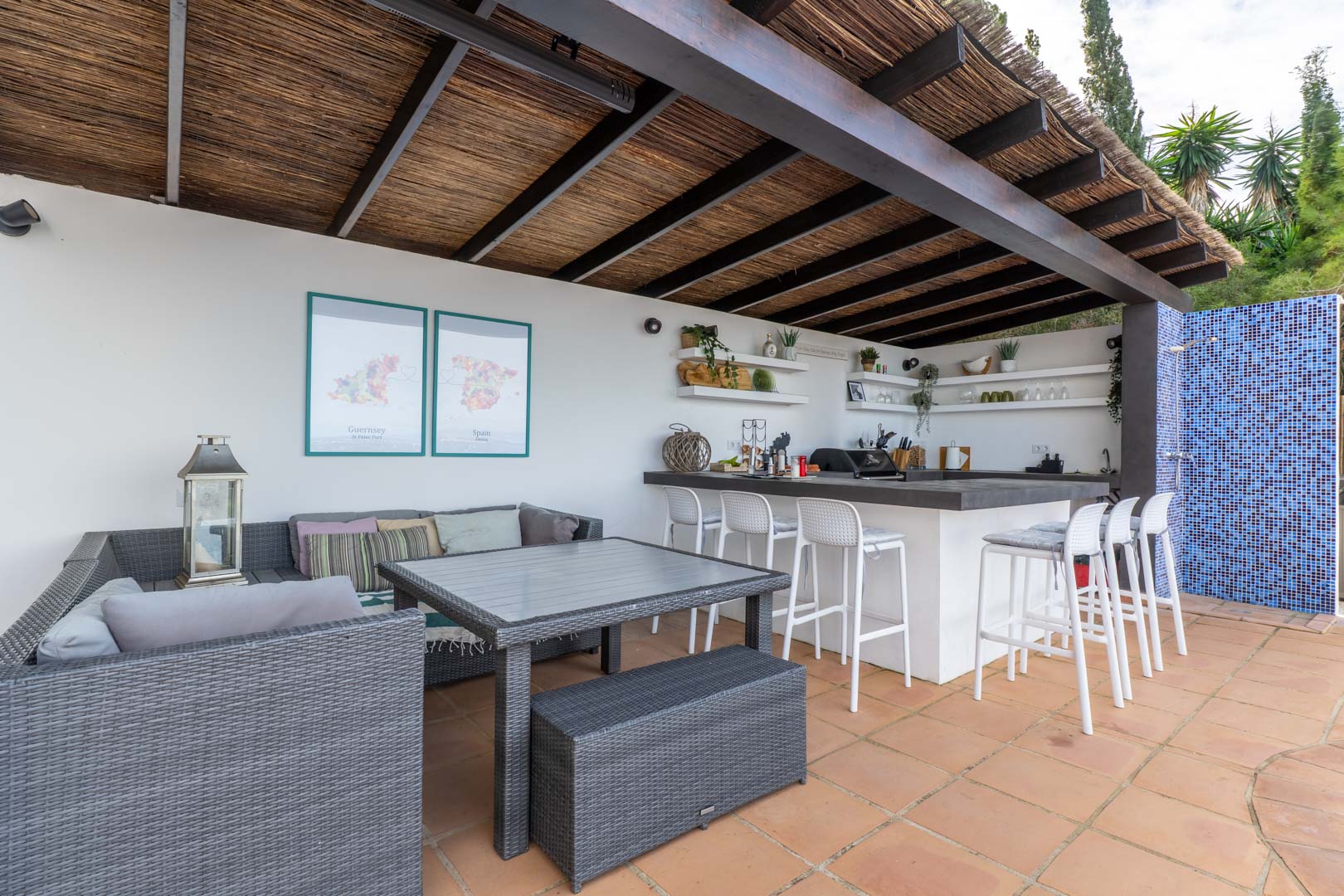 Buitenkeuken en loungegedeelte onder een houten pergola met rieten dak, voorzien van een bar met witte krukken, grijs wicker meubilair en een terracotta vloer.