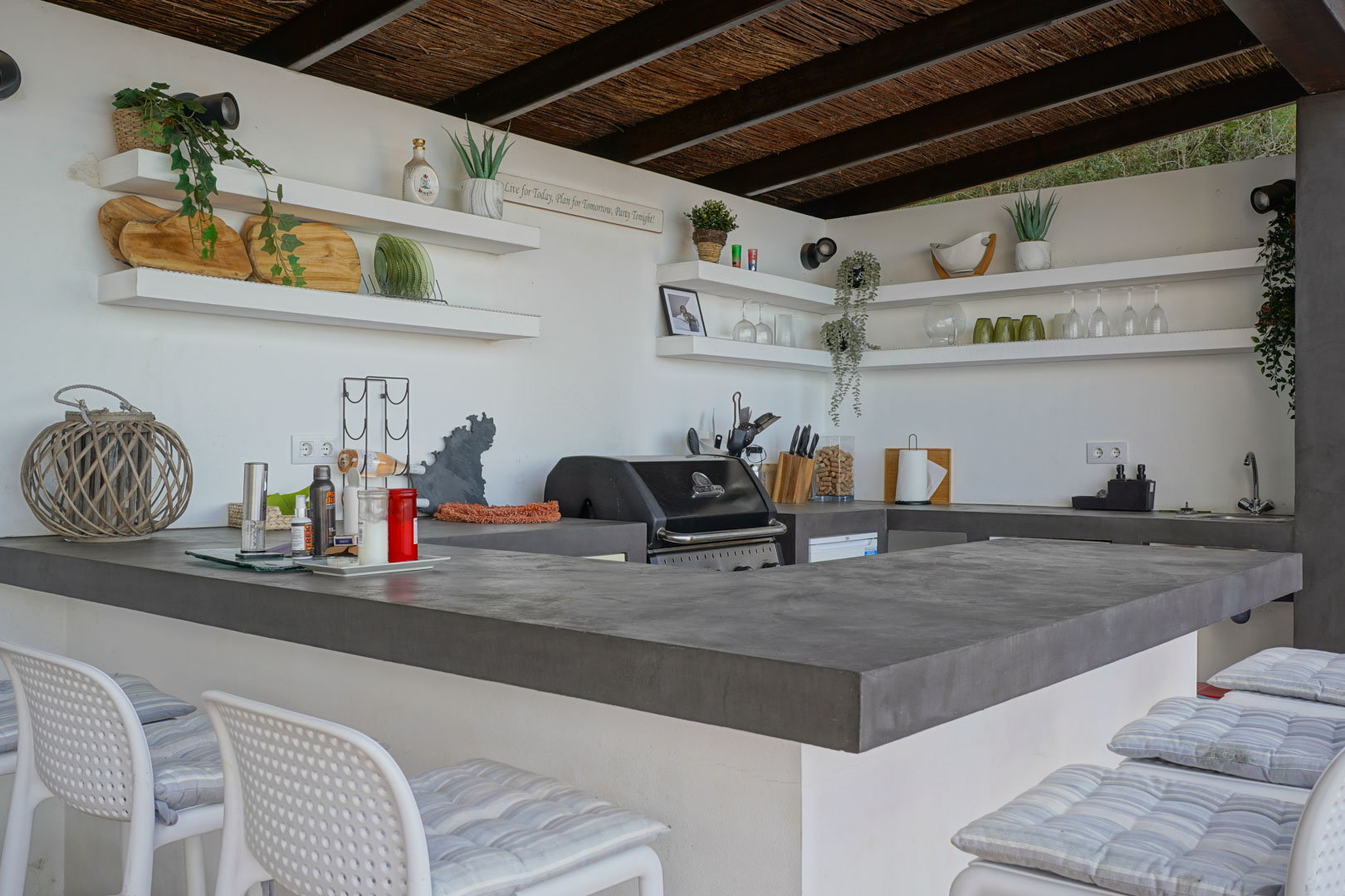 Buitenkeuken met grijze microcement werkbladen, geïntegreerde barbecue, open wandplanken en een rieten plafond met rustieke houten balken.