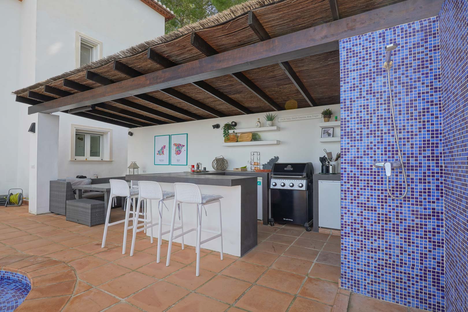 Buitenkeuken onder een houten pergola met rieten dak, voorzien van een gasbarbecue, bar met witte krukken en een buitendouche met blauw mozaïek.