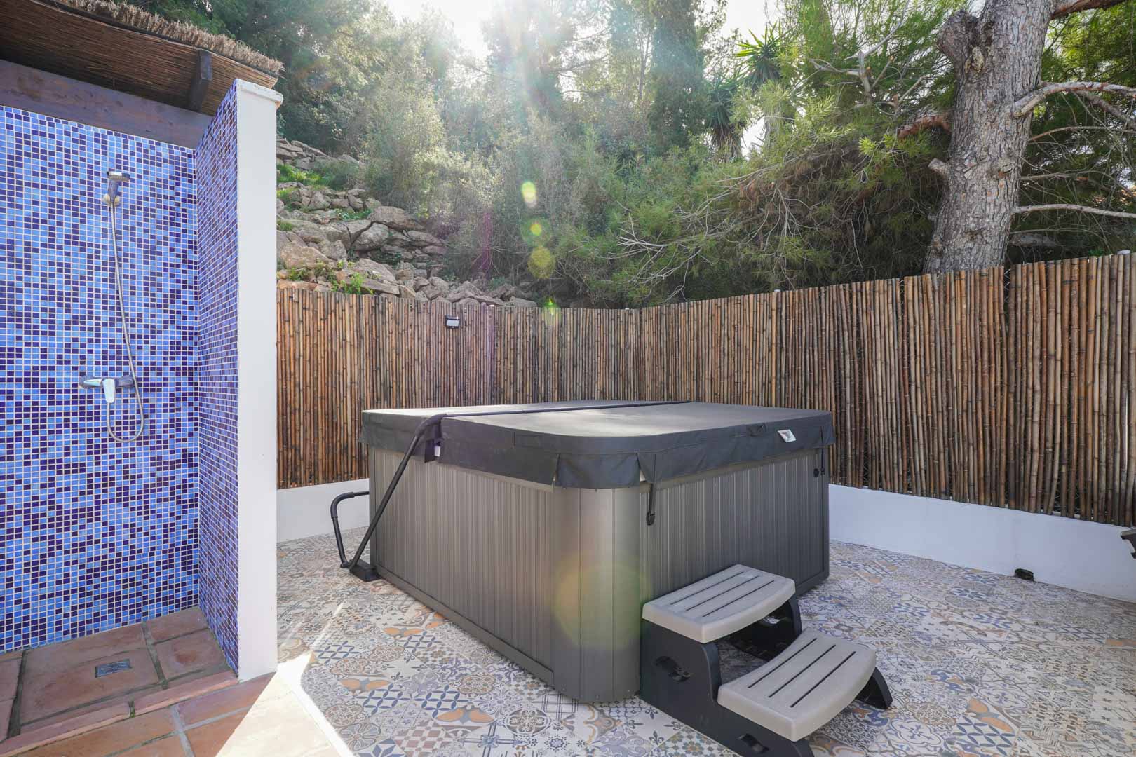 Buitenwellnessruimte met overdekte jacuzzi, blauwe mozaïekdouche en patroontegels, omgeven door een natuurlijke rieten omheining voor optimale privacy.