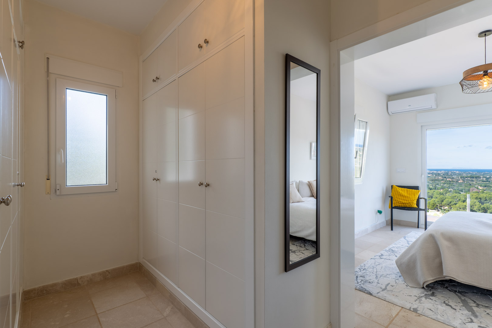 Gang met witte inbouwkasten en passpiegel die toegang biedt tot een slaapkamer met panoramisch zeezicht en airconditioning.