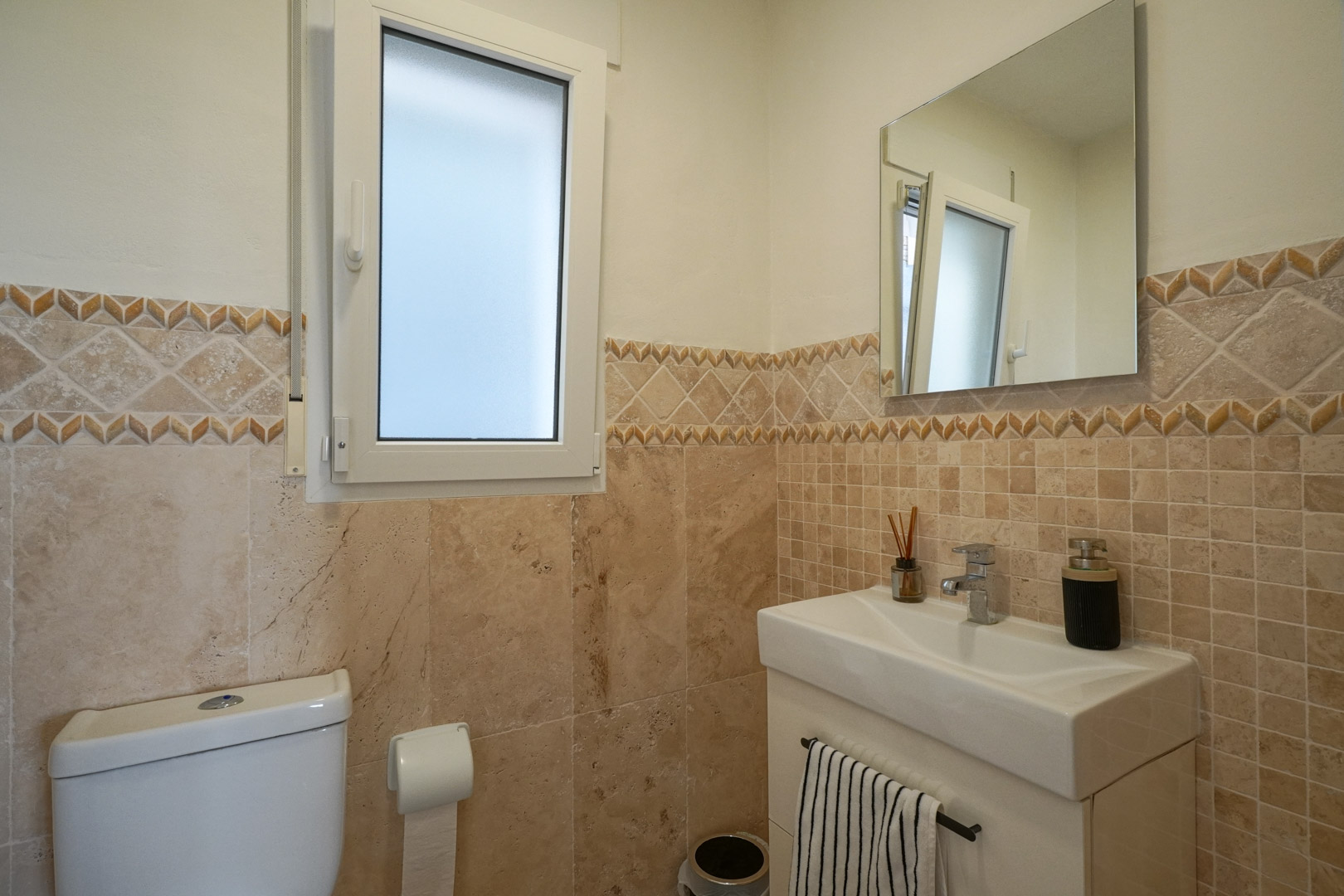 Gastentoilet met aardetinten tegels, decoratieve rand, modern wastafelmeubel en een raam met matglas voor natuurlijke lichtinval.
