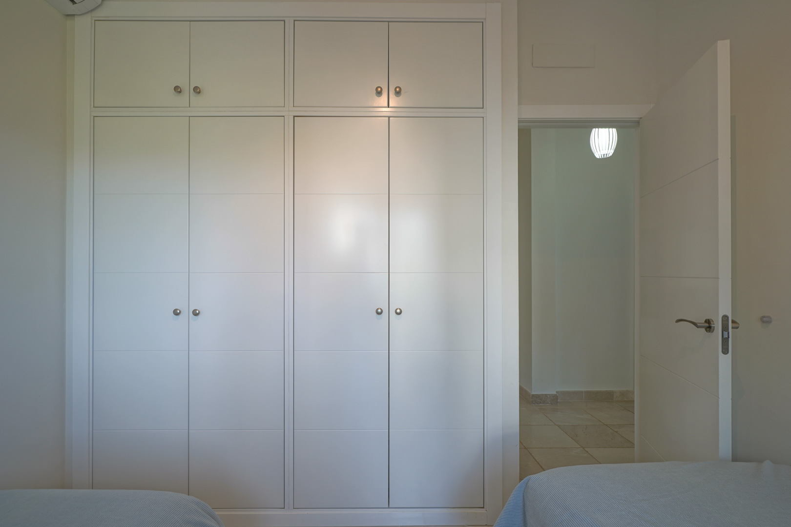 Ingebouwde witte kledingkast met zilveren knoppen en bovenkasten, gesitueerd naast de slaapkamerdeur die uitkomt op een betegelde gang.